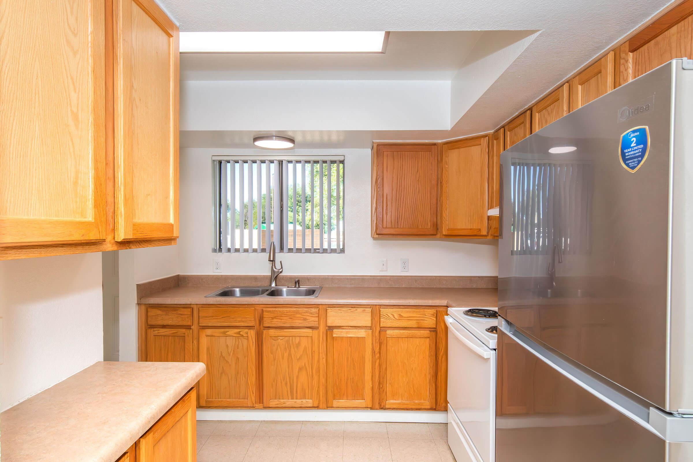 Bright kitchen featuring wooden cabinets, a double sink, and a stainless steel refrigerator. There is a window with vertical blinds allowing natural light to enter, and a modern ceiling light illuminating the space. The countertop is beige and complements the cabinetry.