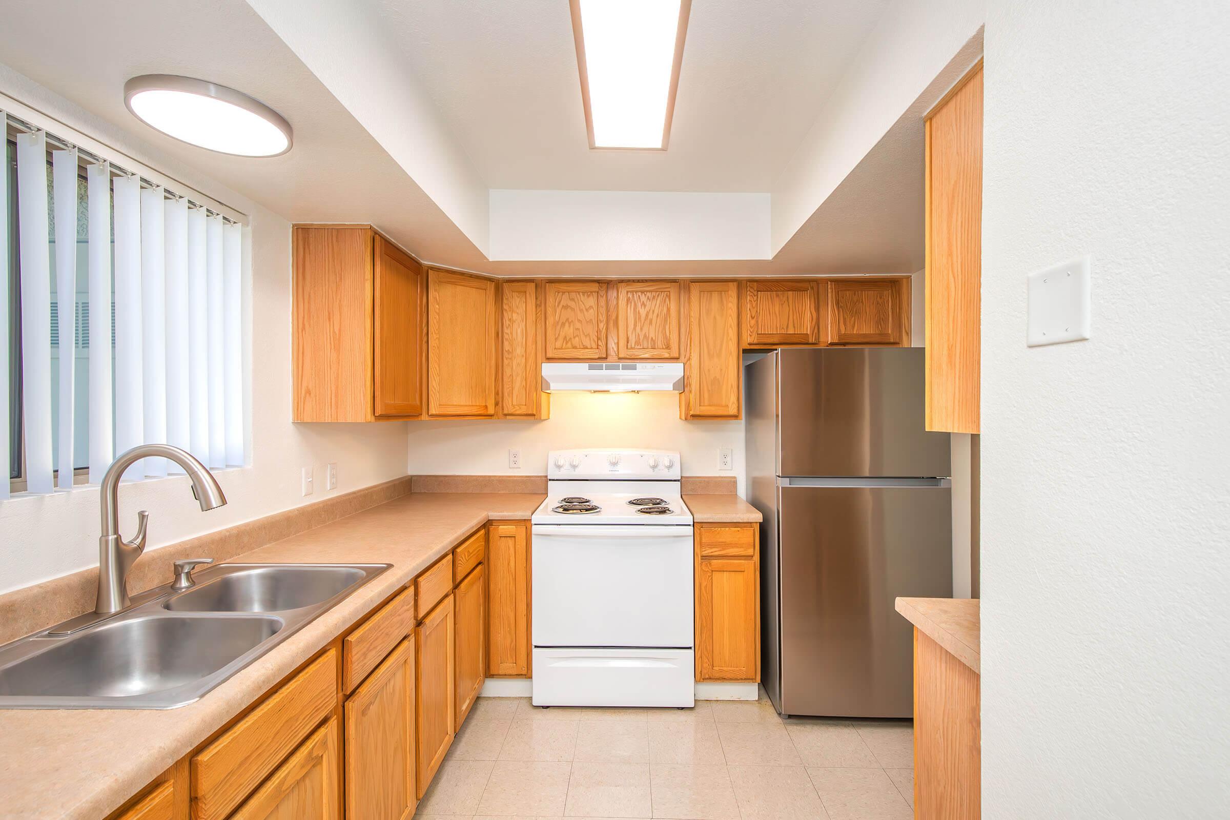 Bright and modern kitchen featuring light wood cabinetry, a stainless steel refrigerator, and a white stove. The countertops are beige, with double sinks beneath a window adorned with vertical blinds. Soft, natural lighting enhances the open and inviting atmosphere of the space.