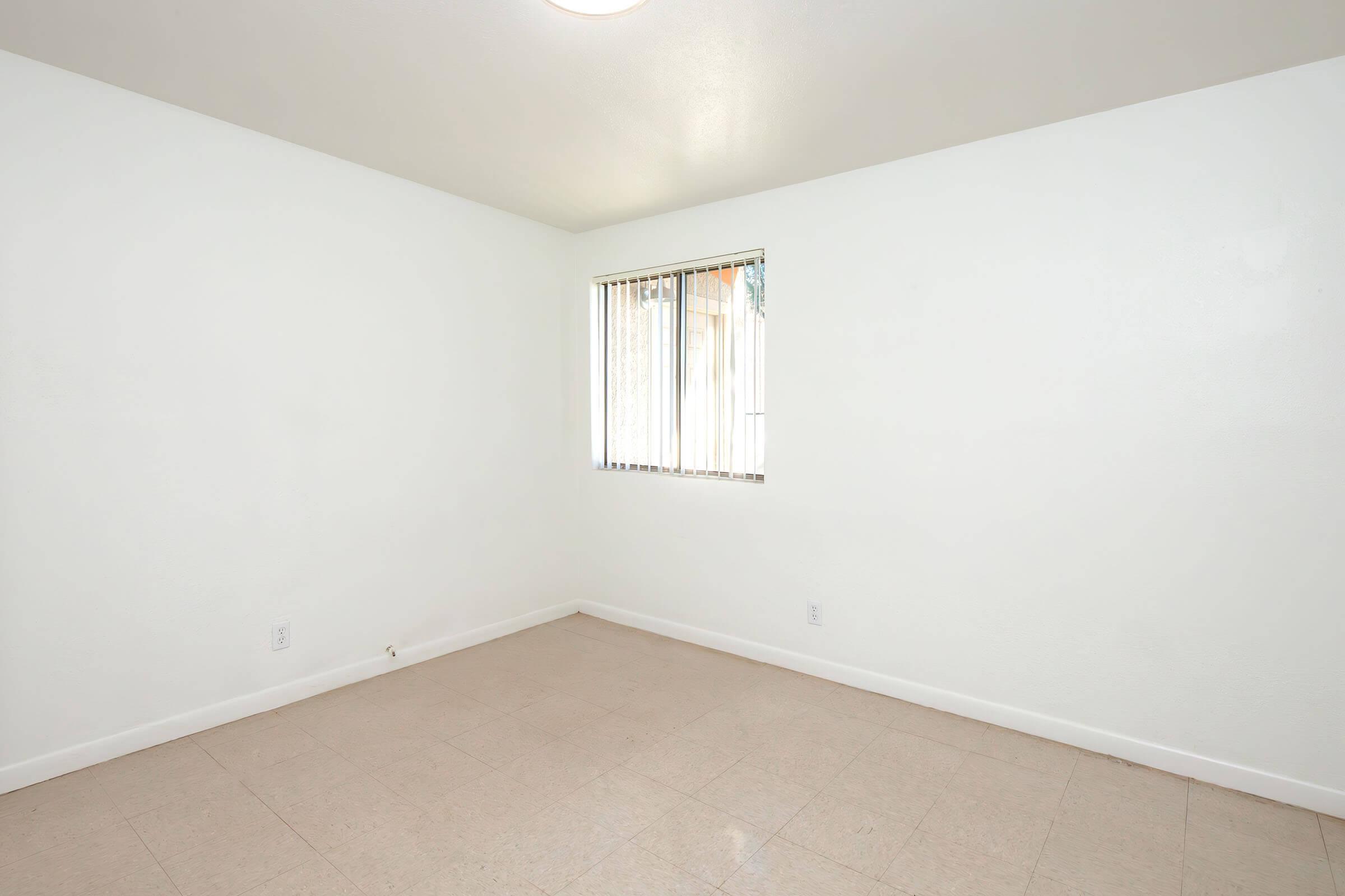 Empty room with light-colored walls and a single window featuring vertical blinds. The floor is tiled with a light-colored material, and the space appears bright and well-lit. No furniture or decorations are present, giving the room a minimalist and clean appearance.