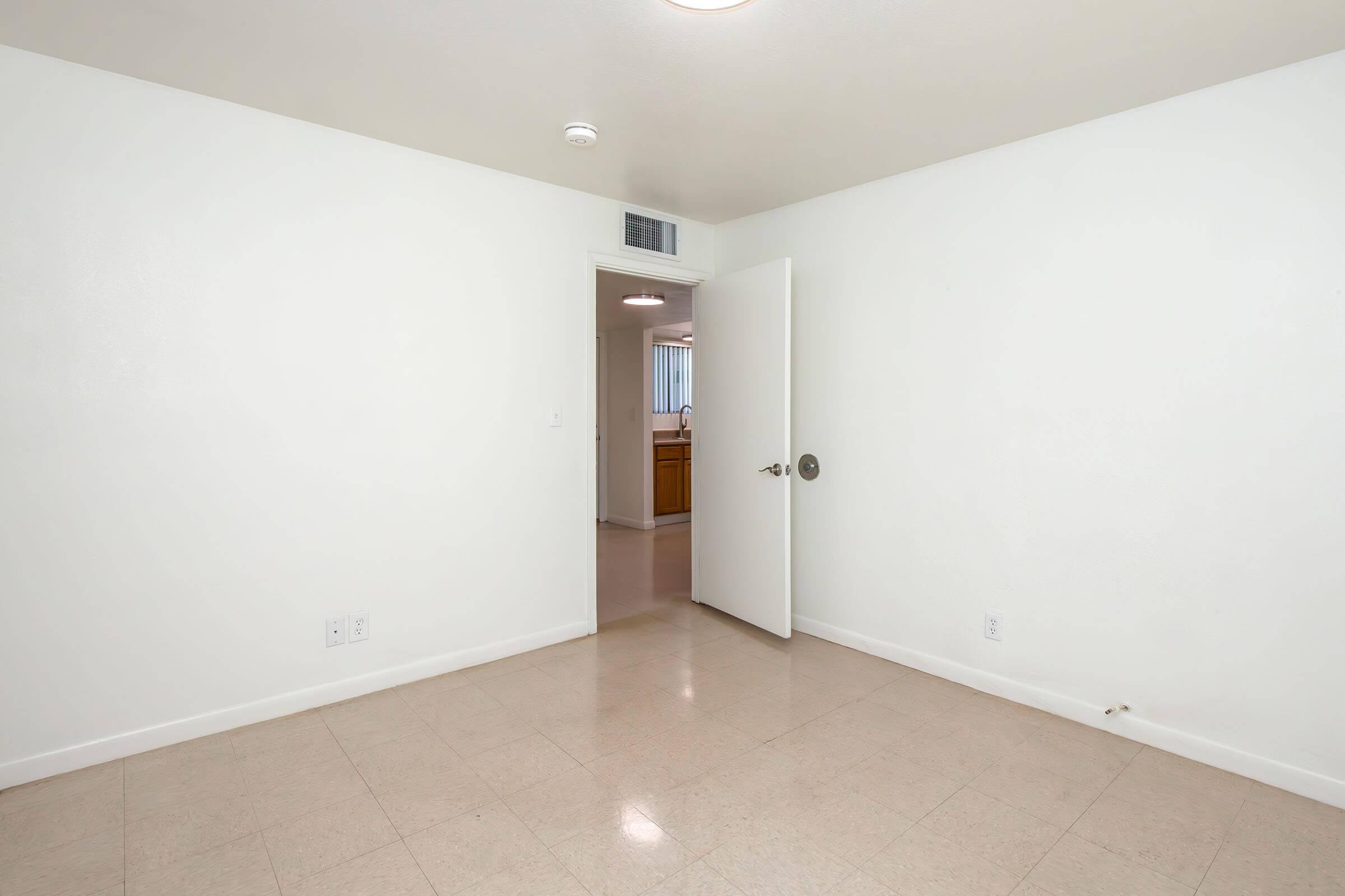 Empty room with white walls, beige tile flooring, and an open door leading to another room. The interior is well-lit and unadorned, creating a spacious and minimalist ambiance. 