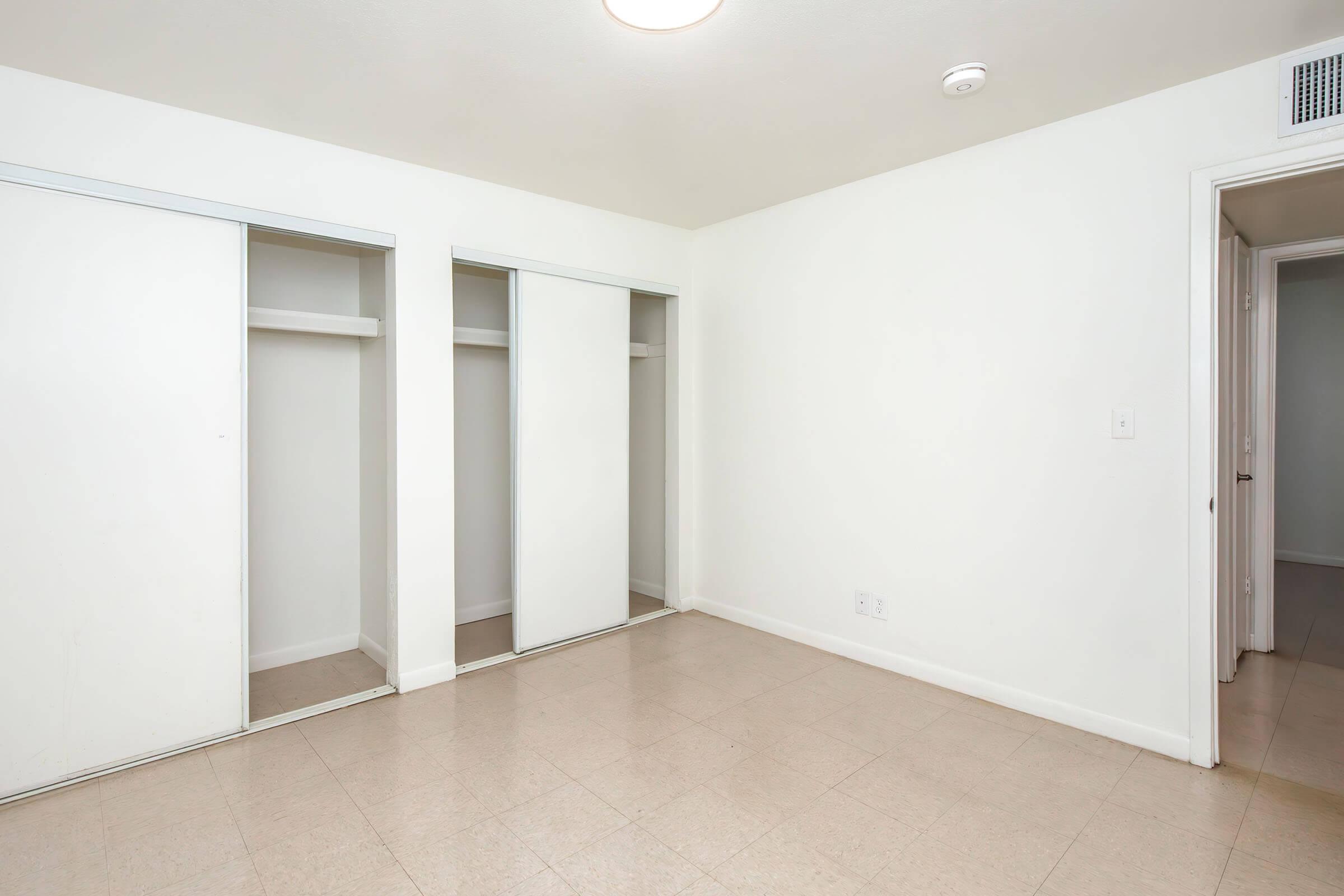 Empty room with white walls and a single ceiling light. It features two sliding closet doors to the left and a doorway leading to another room on the right. The floor is tiled in a light color, creating a clean and simple appearance.