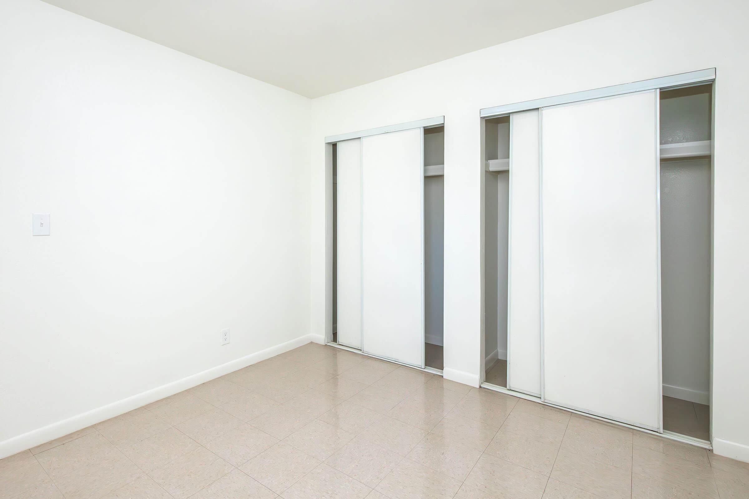 A minimal, empty room featuring two sliding door closets against a plain white wall. The floor has a light-colored tile, and the overall ambiance is bright and uncluttered, suggesting a blank canvas for potential furnishing and decoration.
