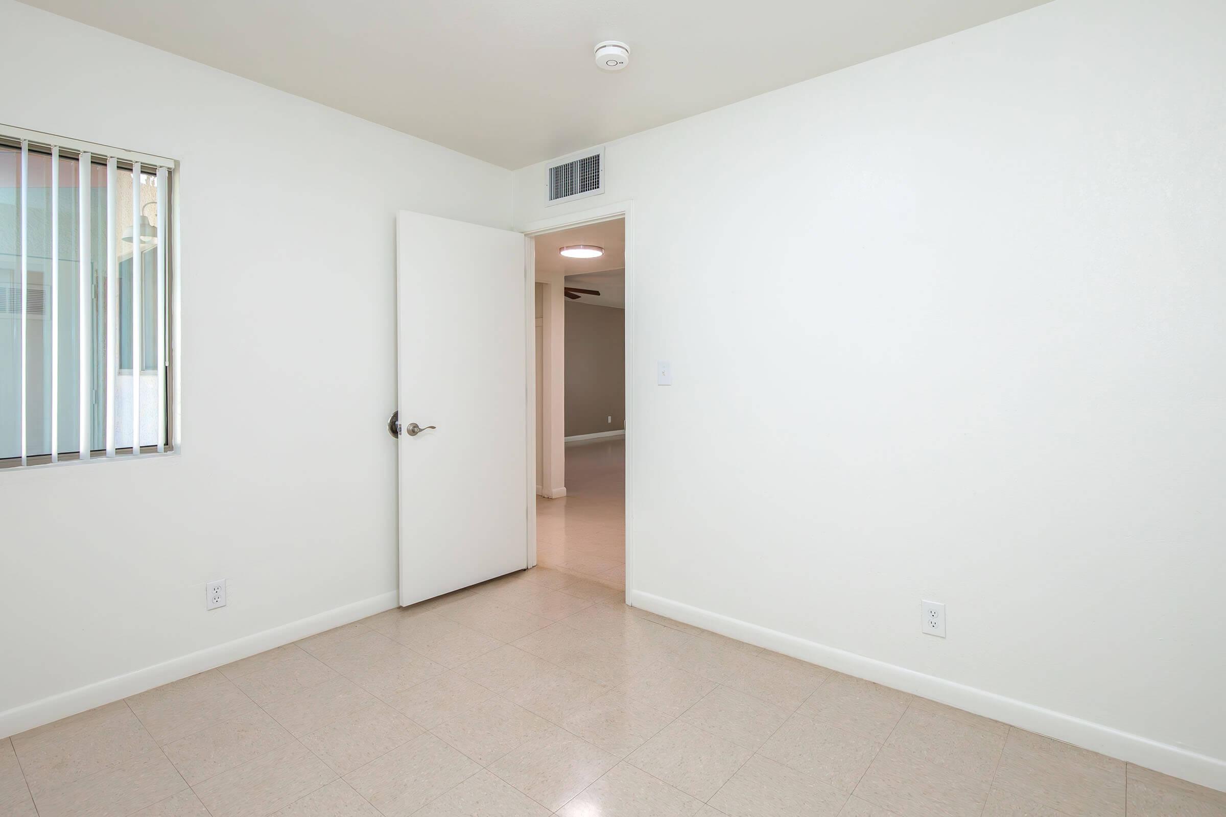 A bright, empty room with white walls and tiled flooring. There is a door on the right, leading to another room, and a window with blinds on the left. The space is clean and minimalistic, offering a sense of openness.