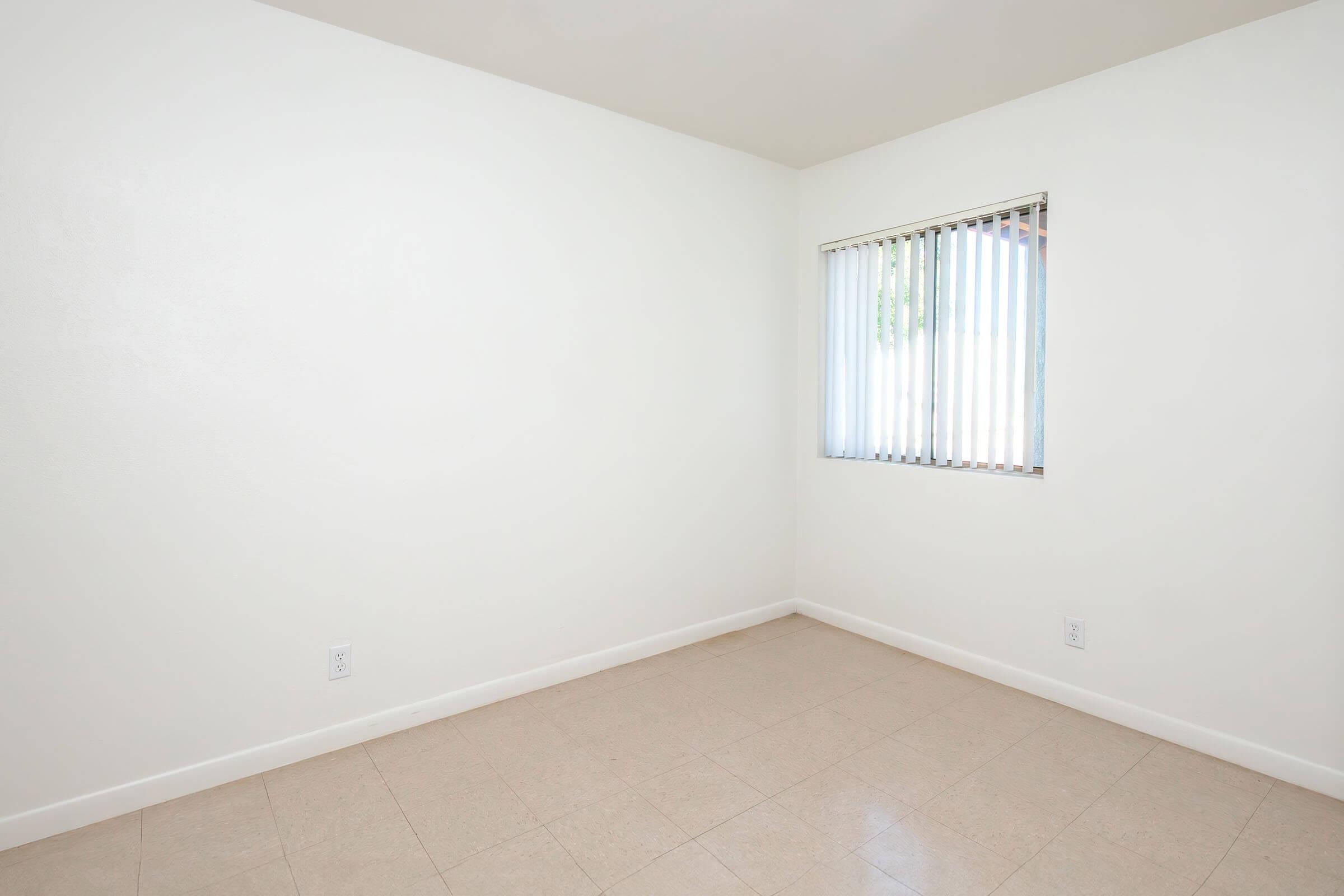 Empty room with light-colored walls and a light tile floor. A window with vertical blinds is visible on one wall, allowing natural light to enter the space. The room is unfurnished and has a clean, minimalistic appearance.