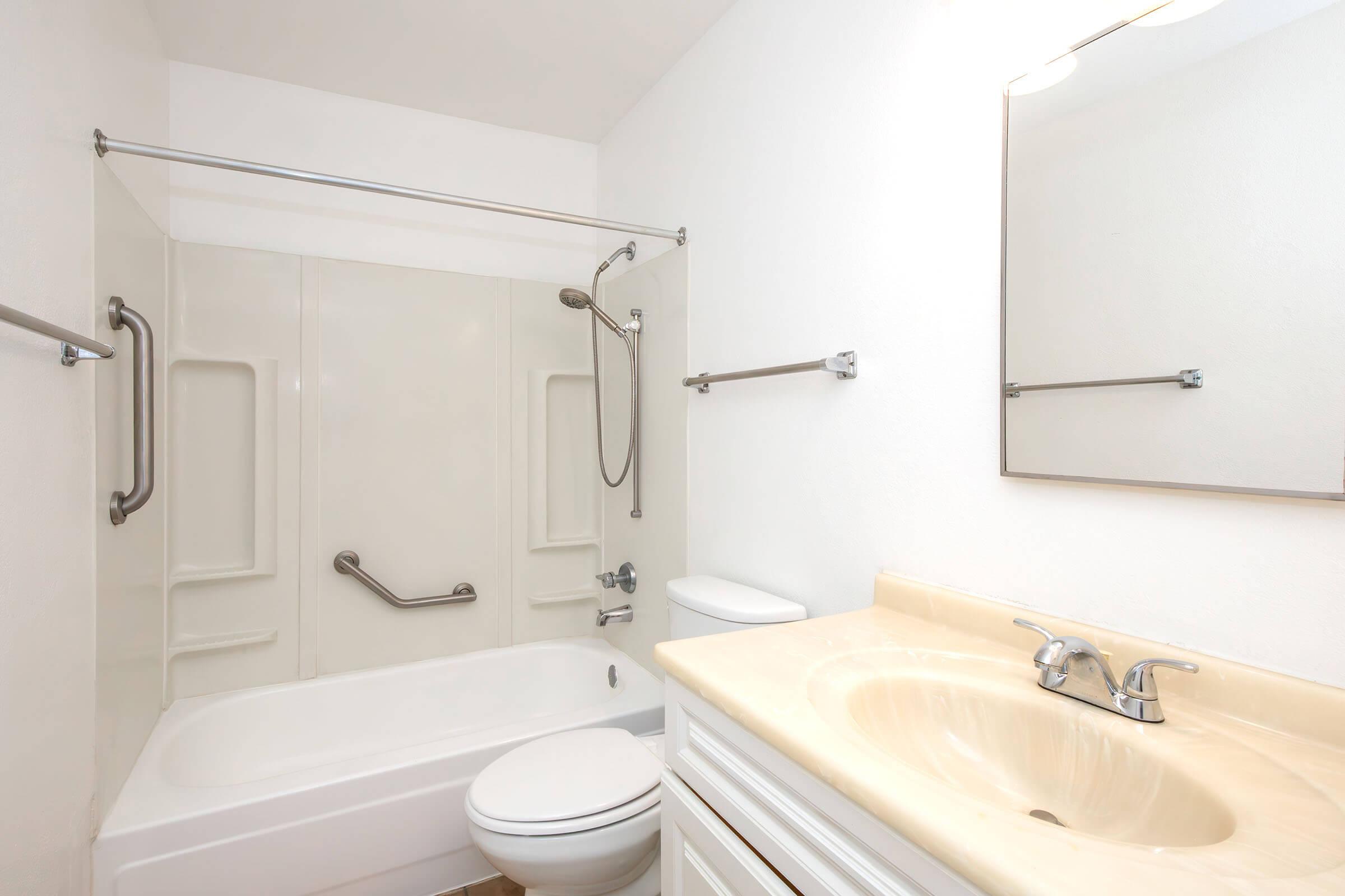 A clean and modern bathroom featuring a bathtub with a shower, grab bars for safety, a white toilet, and a sink with a beige countertop. The walls are light-colored, and there is a mirror above the sink, providing a bright and spacious appearance.