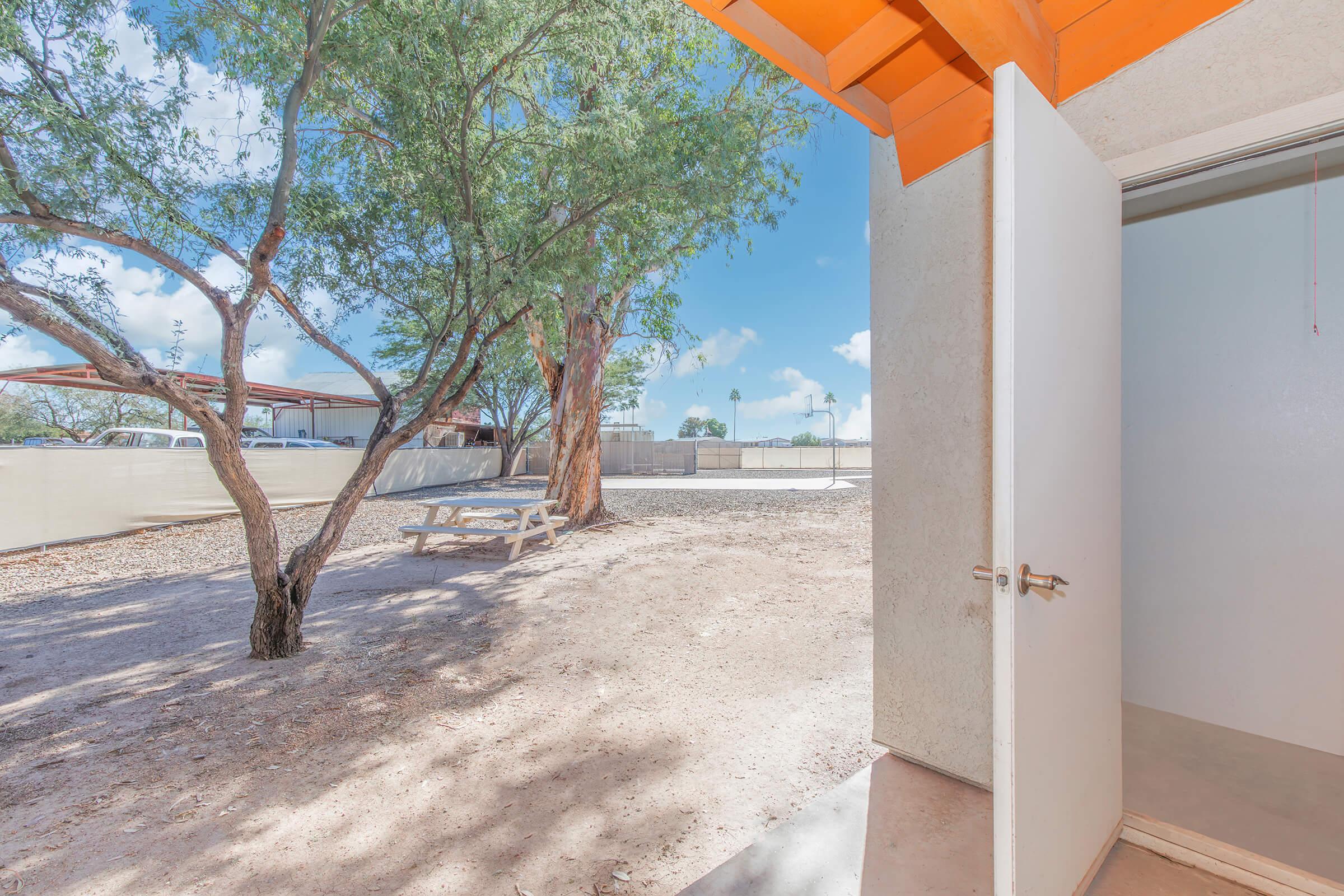 A view from an open door showcasing a dry landscape with a large tree and a picnic table nearby. The sky is bright with scattered clouds, and there are structures in the background. The scene conveys a welcoming, outdoor space.