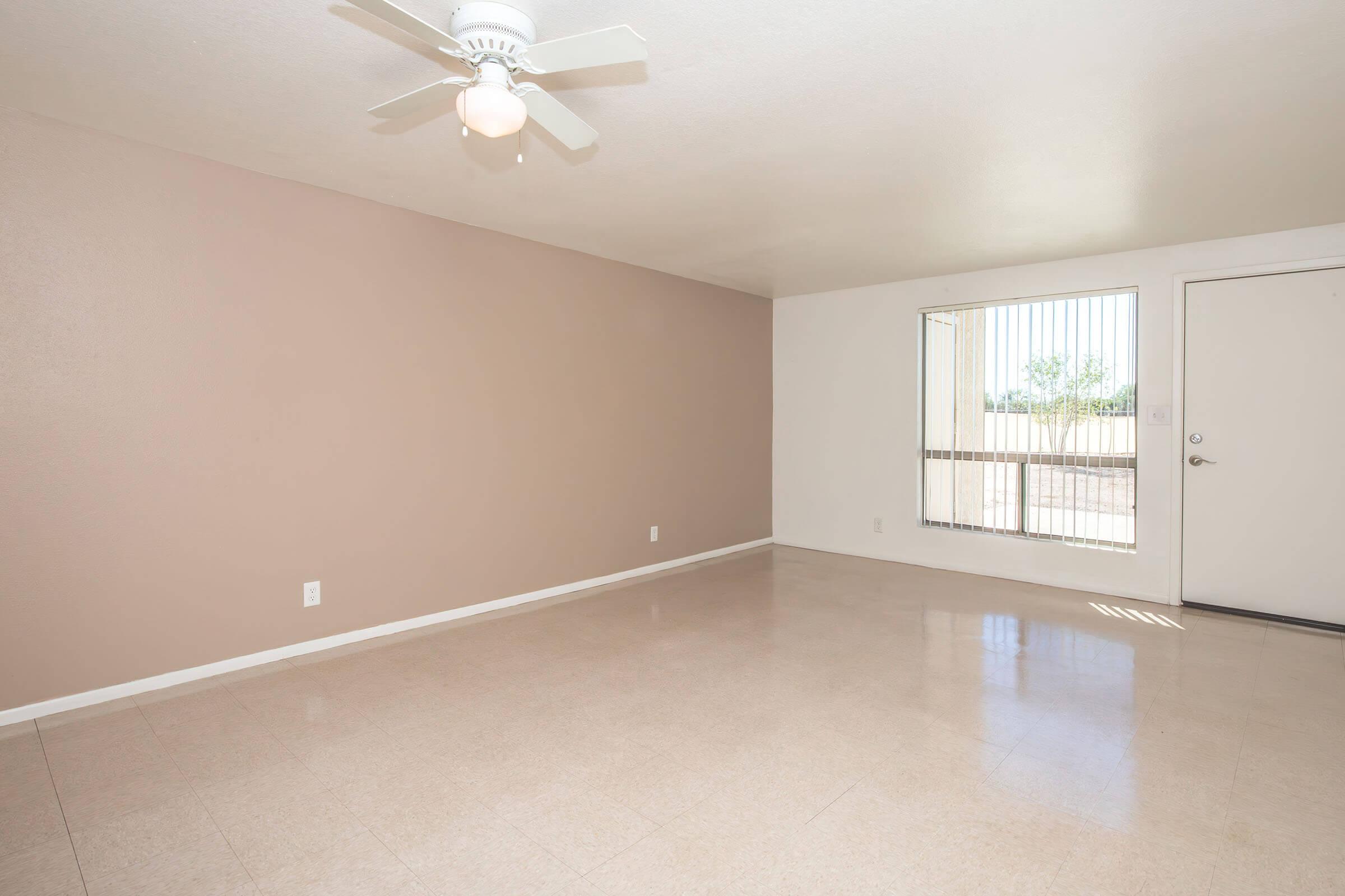 A spacious and empty living room featuring a light-colored ceiling fan, beige wall, white wall, and large window with blinds. The floor is tiled with a glossy finish, and there is a door leading outside. Natural light brightens the room, creating an open and airy atmosphere.