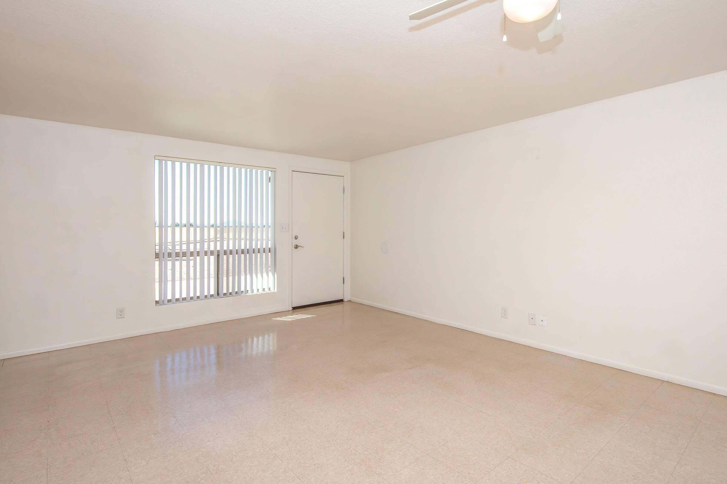 Empty room with a light-colored floor and white walls. There is a window with vertical blinds on the left and a door on the right. The ceiling features a ceiling fan. The space is well-lit and has no furniture, creating a minimalist and open environment.