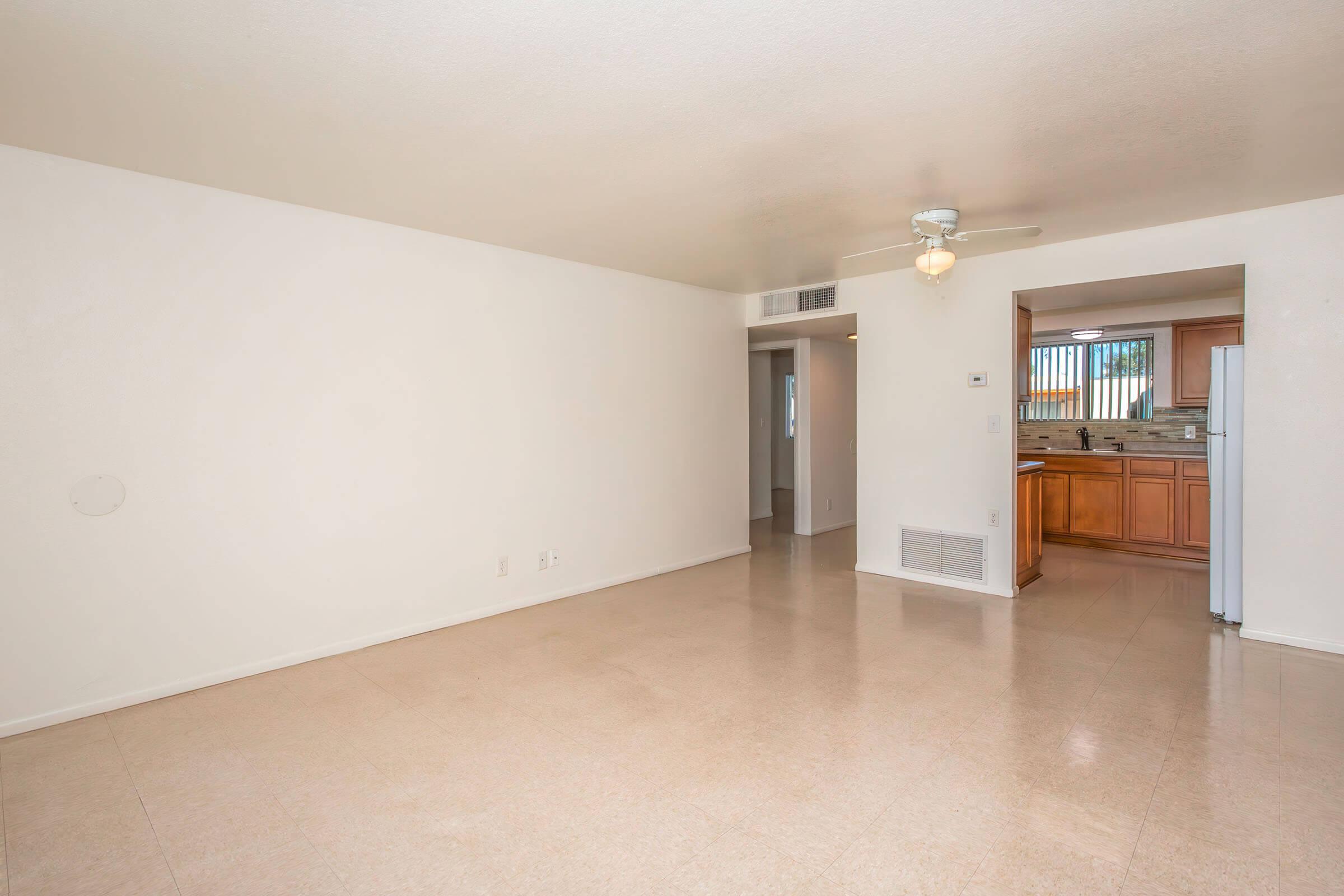 A spacious, empty living room with light-colored walls and a ceiling fan. The floor is tiled in a light shade, and there is a doorway leading to a kitchen visible in the background. Sunlight enters through a window, creating a bright and airy atmosphere.