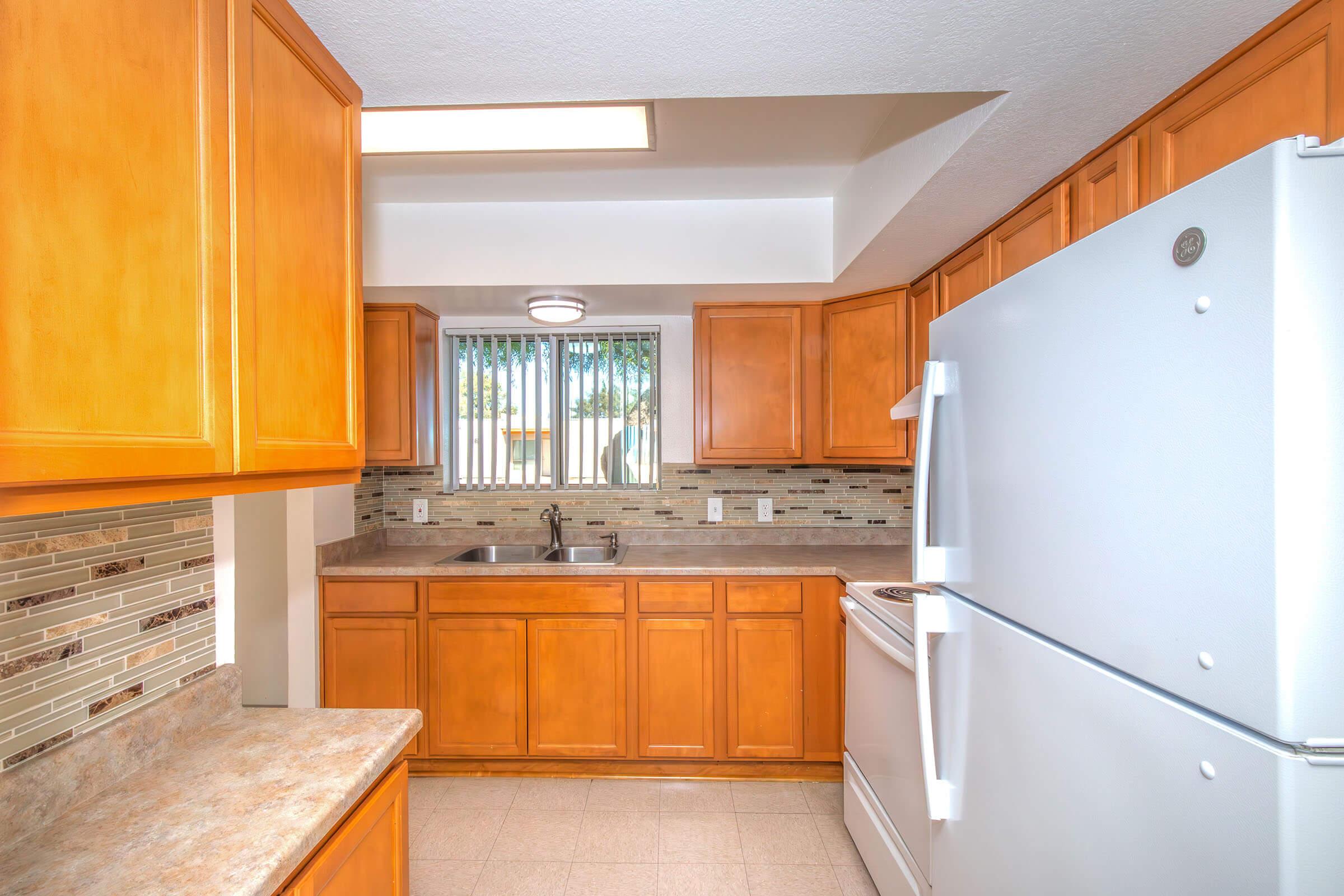 Bright kitchen featuring wooden cabinets, a stainless steel sink under a window, and modern appliances including a white refrigerator and stove. The countertop is light-colored, and the walls are decorated with a stylish backsplash of mixed tiles.