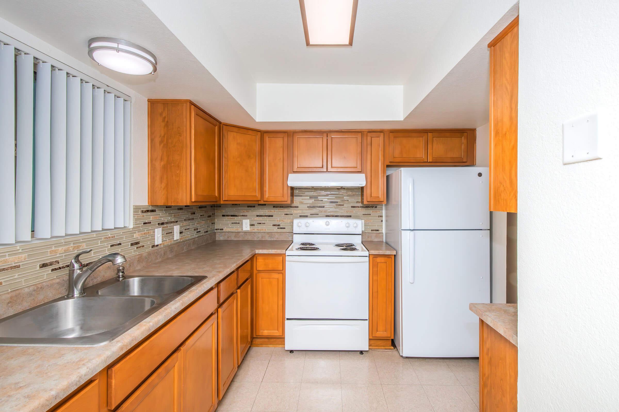 A modern kitchen featuring wooden cabinets, a double sink, a white gas stove, and a white refrigerator. The walls are adorned with light-colored tile backsplash, and there are blinds on the window. The space is brightly lit with a ceiling light.