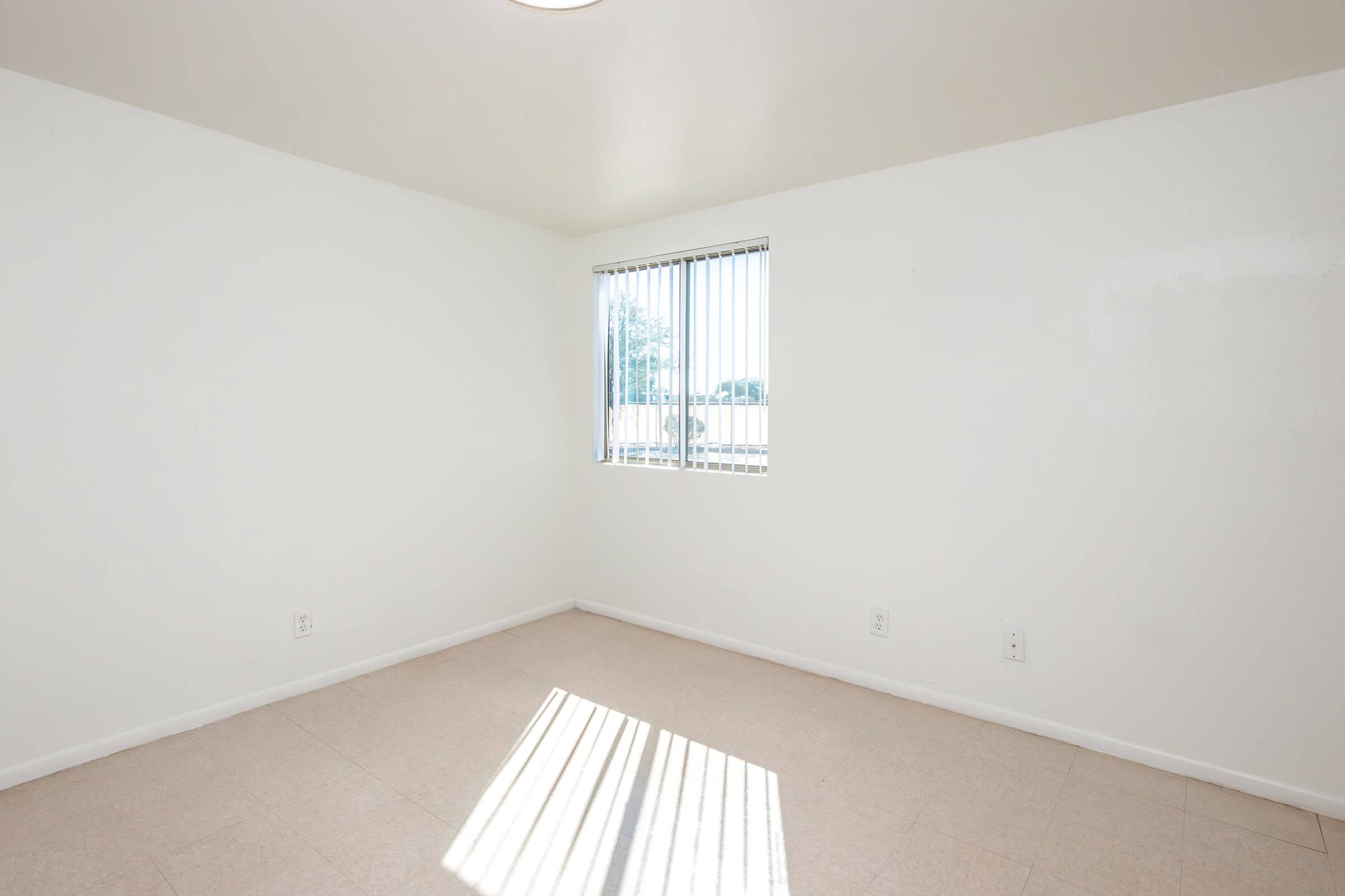 Empty room with beige floor tiles, white walls, and a window with vertical blinds allowing sunlight to cast stripes on the floor. The space is minimalistic and bright, suggesting potential for furniture placement or decoration.