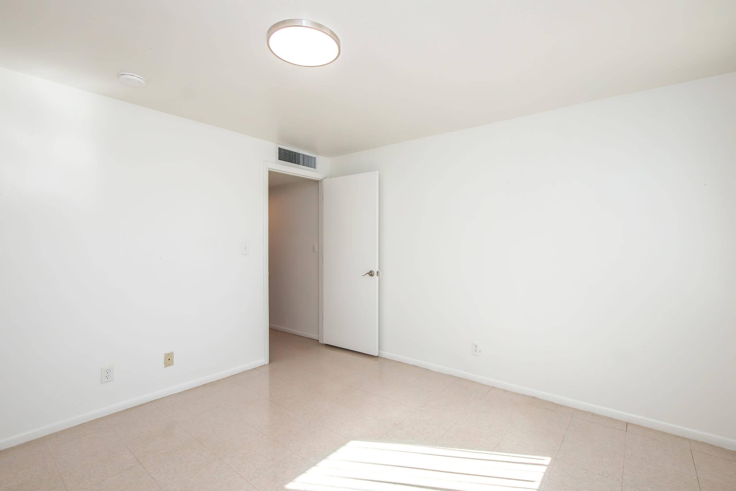 A simple, empty room with white walls and a light-colored floor. The ceiling features a round light fixture, and there is a closed door on the right leading to another space. Sunlight casts a shadow on the floor, indicating a bright atmosphere. The room feels spacious and clean, ready for furnishings.