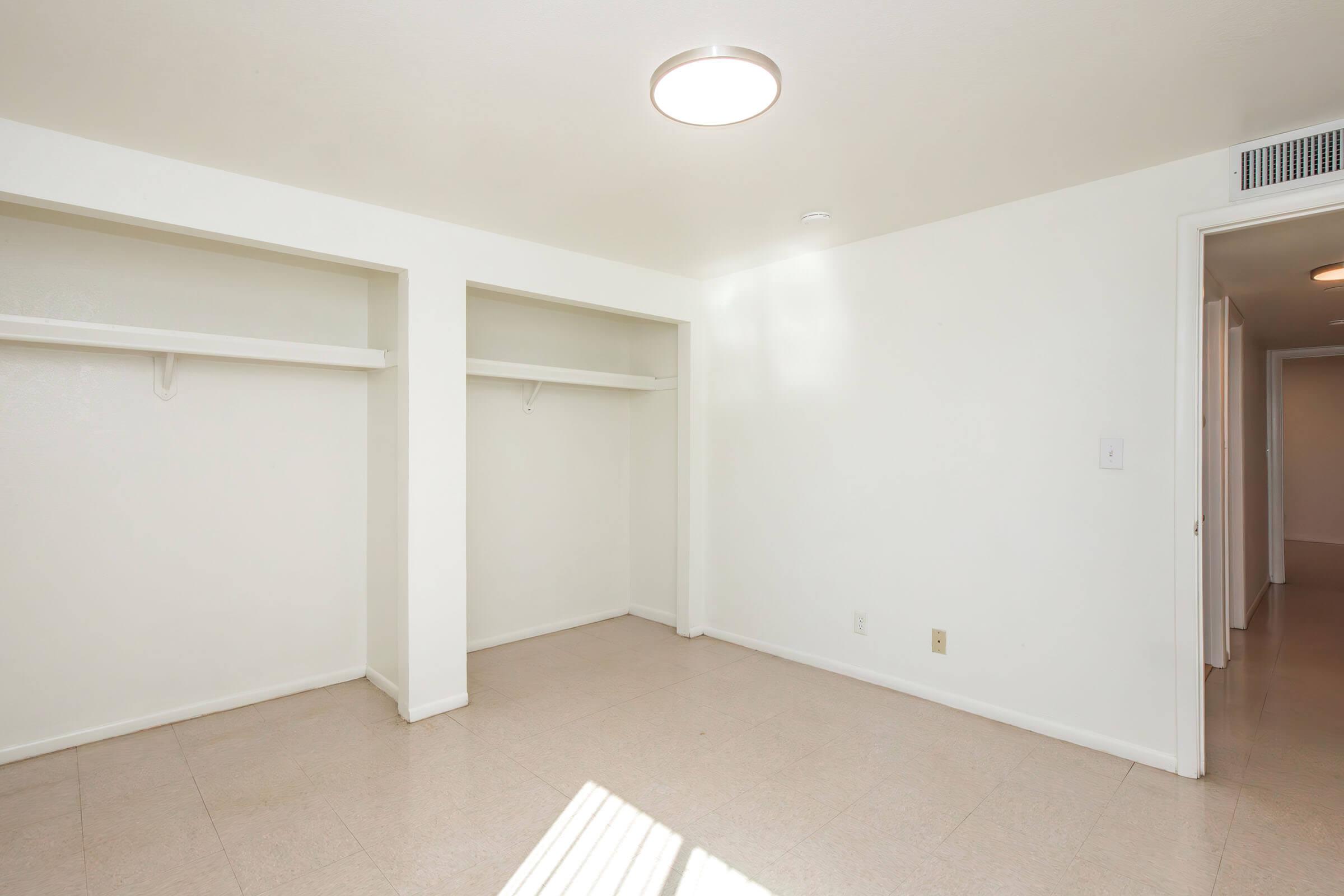 A spacious, empty room with light-colored walls and a light fixture on the ceiling. The room features two closet openings on the left side and a doorway on the right leading to another area. Natural light illuminates the space, highlighting the clean, minimalistic design and beige tiled floor.