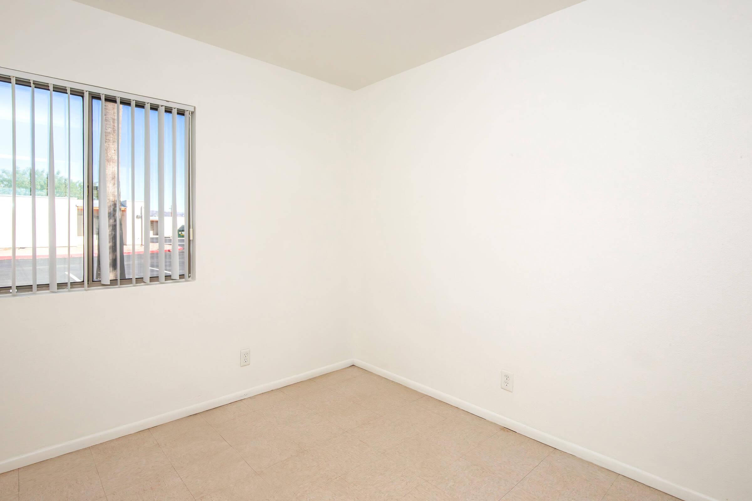 Empty room with white walls and light-colored flooring. A window with vertical blinds allows natural light to enter, casting a soft illumination. The space appears clean and unfurnished, highlighting its simplicity and potential for various uses.