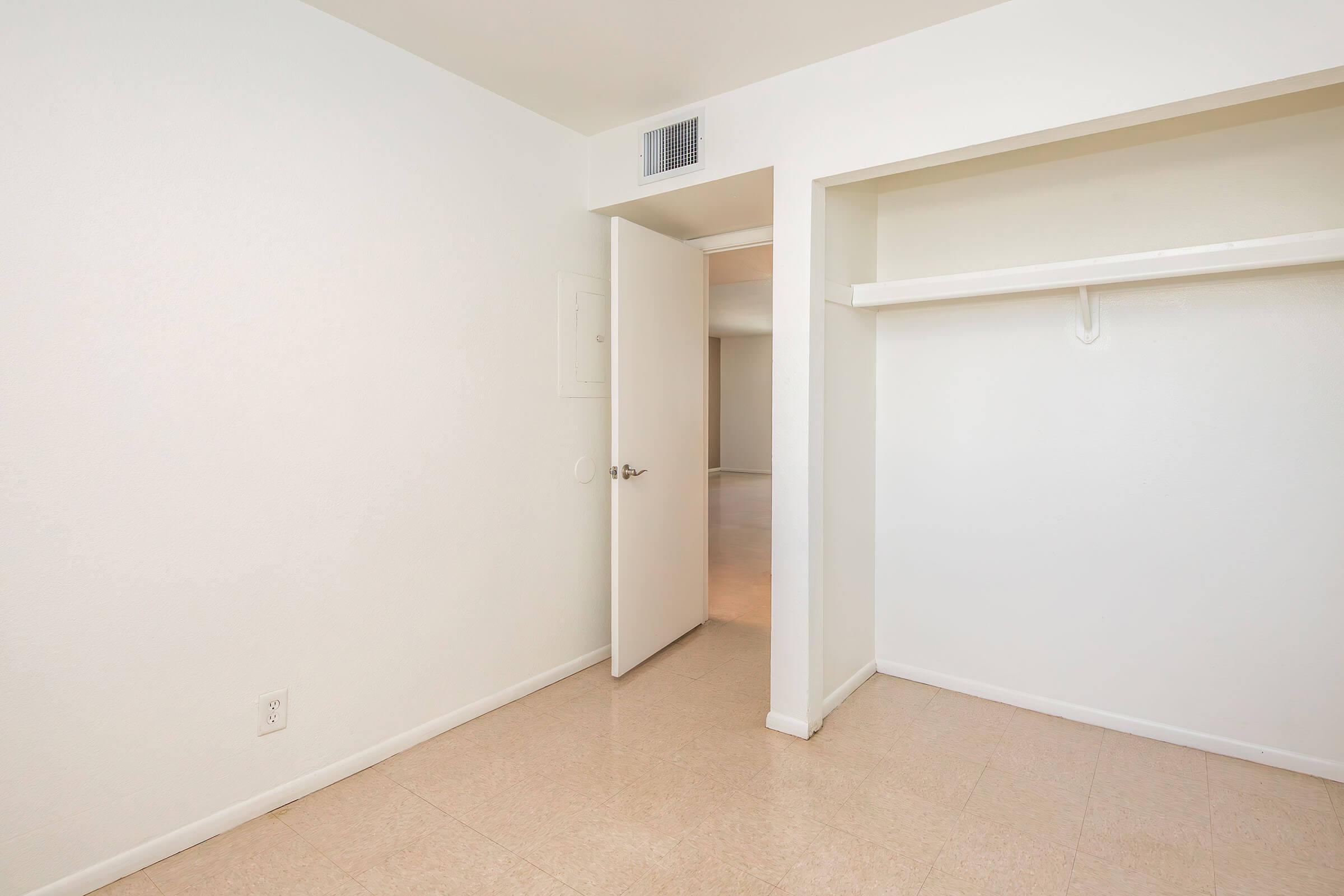 A small, simple room with white walls and a light-colored floor. There is an open door leading to another room, and a closet space with a shelf against one wall. The room is well-lit and has a clean, minimalist appearance.