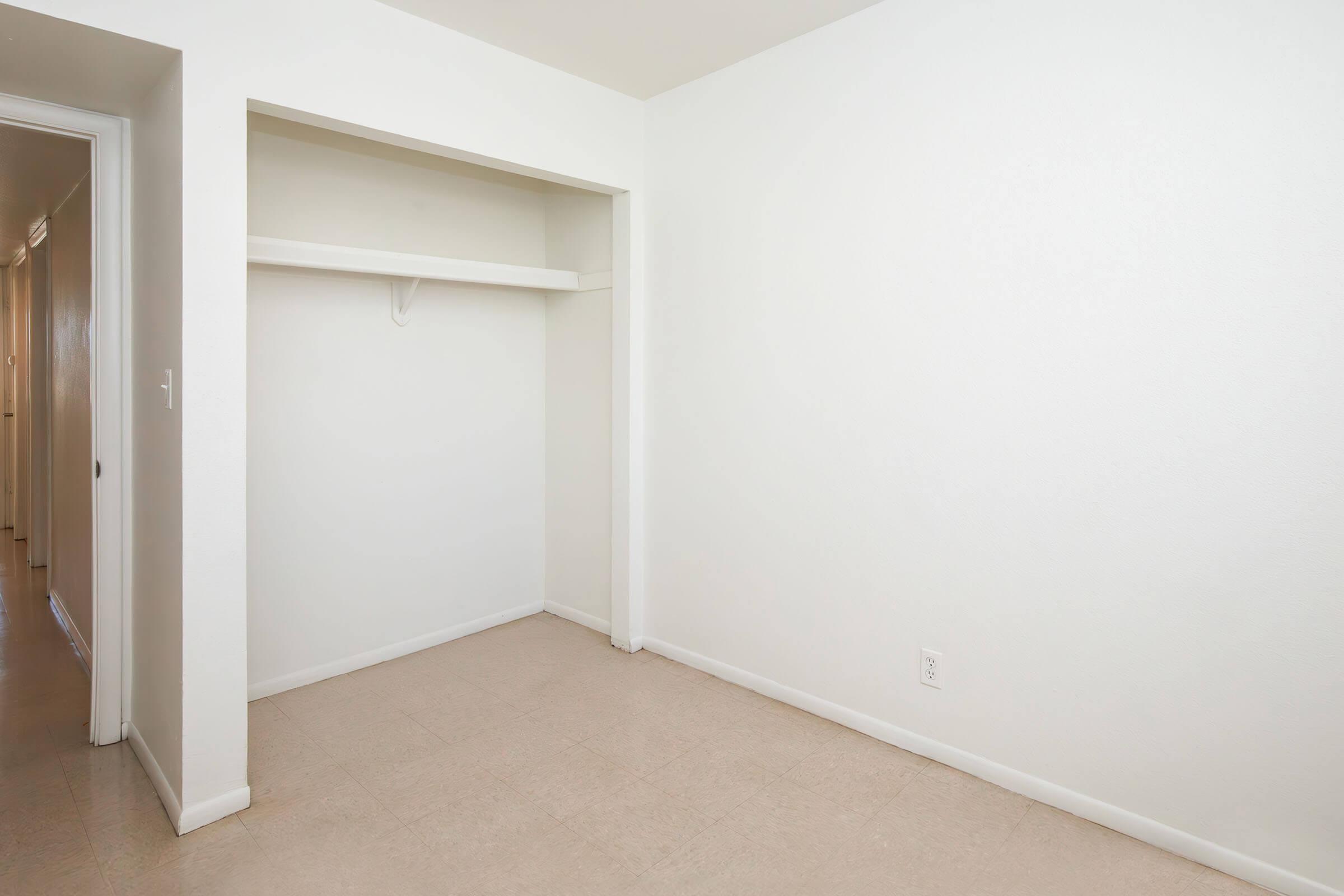 A vacant room with white walls and a light-colored floor. There is an empty closet on one side with a shelf, and the other side features a doorway leading to a hall. The space appears bright and minimally furnished, creating a neutral and open atmosphere.