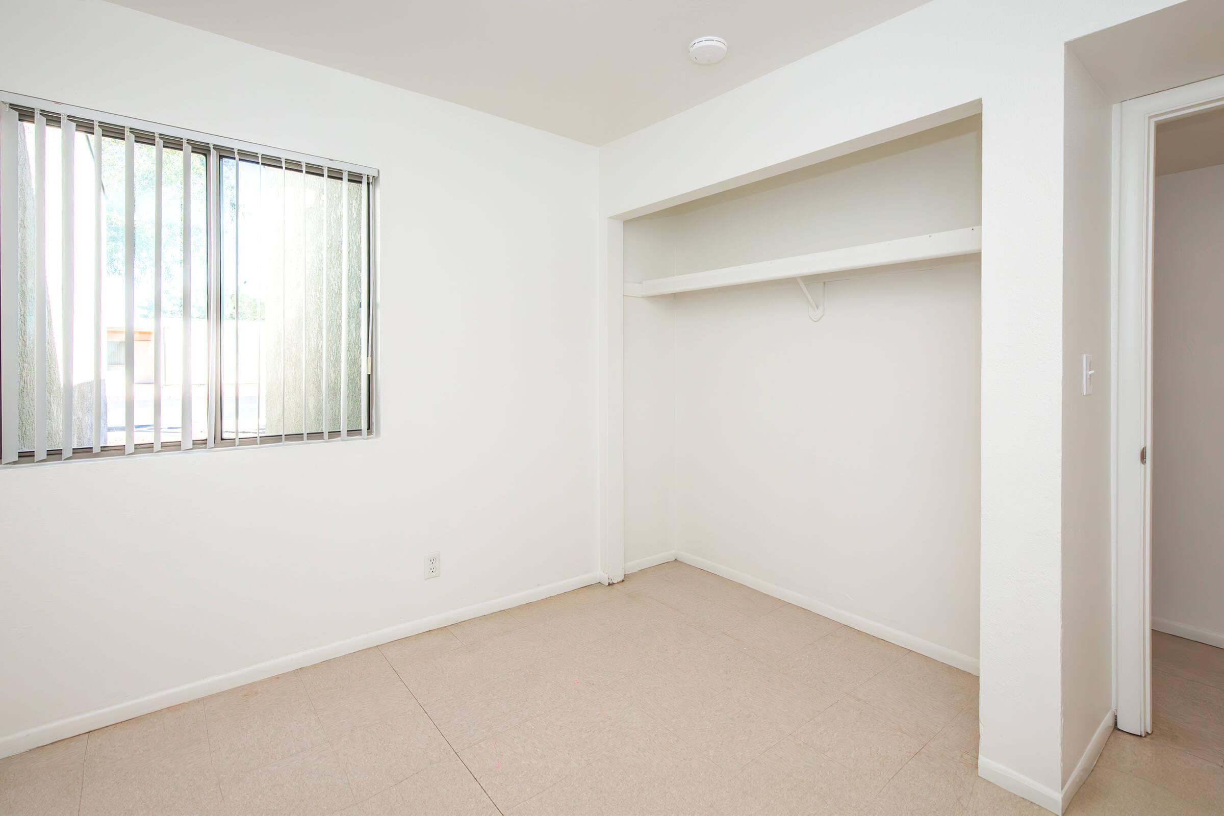 A vacant room with white walls and a light-colored floor. There is a window with vertical blinds allowing natural light in, and a closet space on the left without doors. The overall atmosphere is clean and minimalistic, suitable for personal decoration or use.