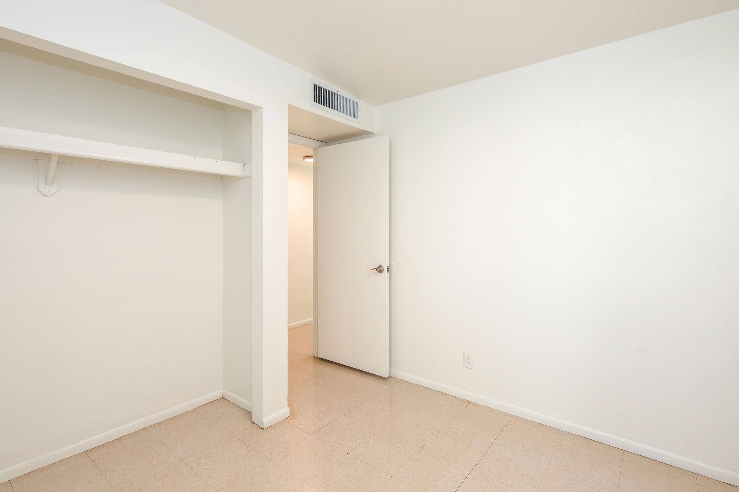 Empty room with white walls and a tiled floor. There is a closet on the left with a shelf and an open door leading to another space. The lighting is soft and the overall ambiance is minimalistic and clean.