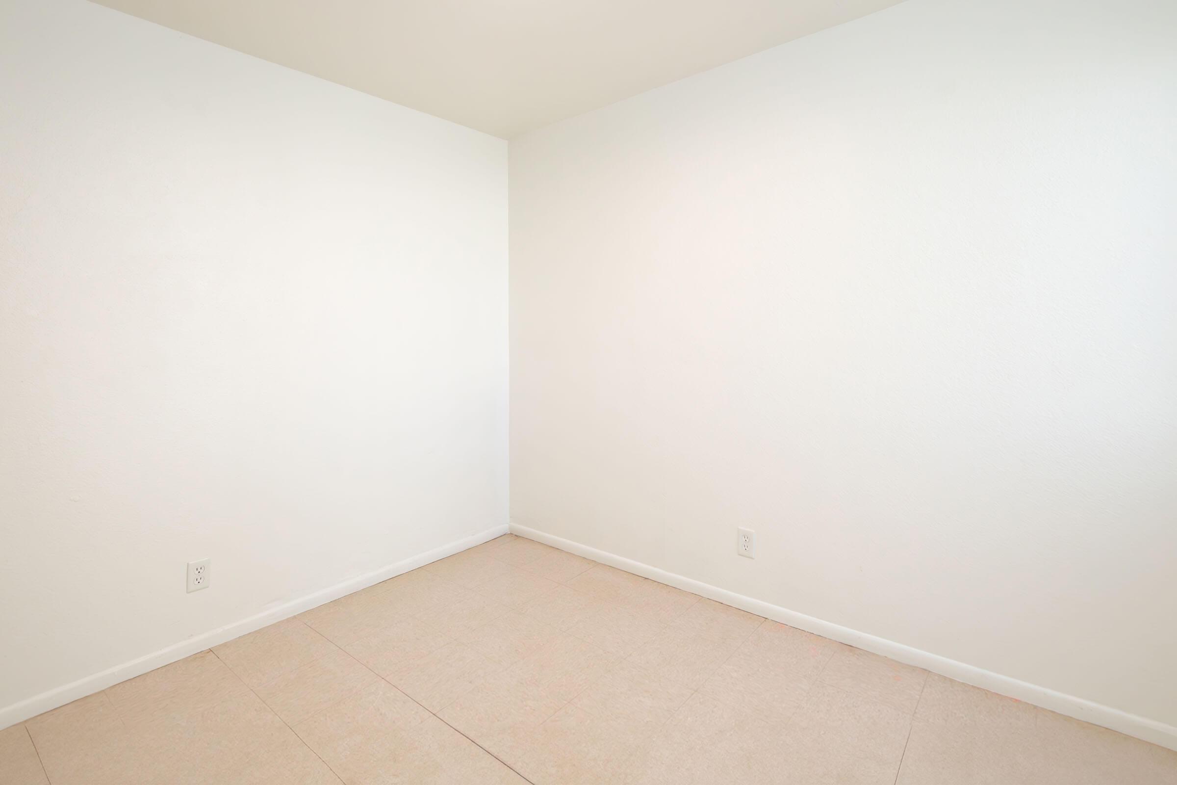 Empty room with plain white walls and a light-colored floor. There are no furnishings or decorations, creating a minimalist and spacious appearance. The lack of windows or features emphasizes the simplicity of the space.