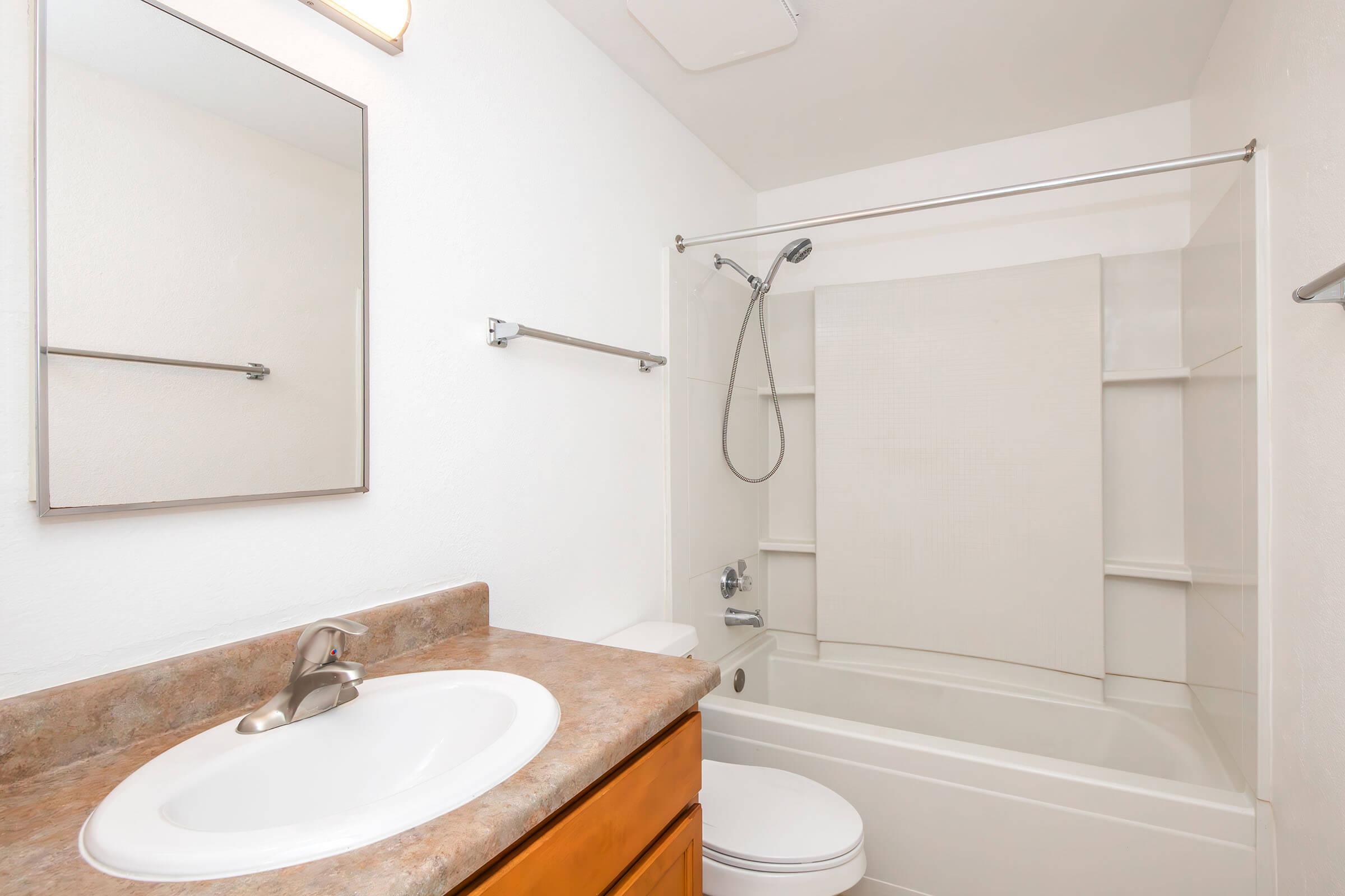 A clean and modern bathroom featuring a bathtub with a shower, a white wall, a mirror above a sink with a countertop, and simple fixtures. The space is well-lit and tidy, highlighting a minimalist design.