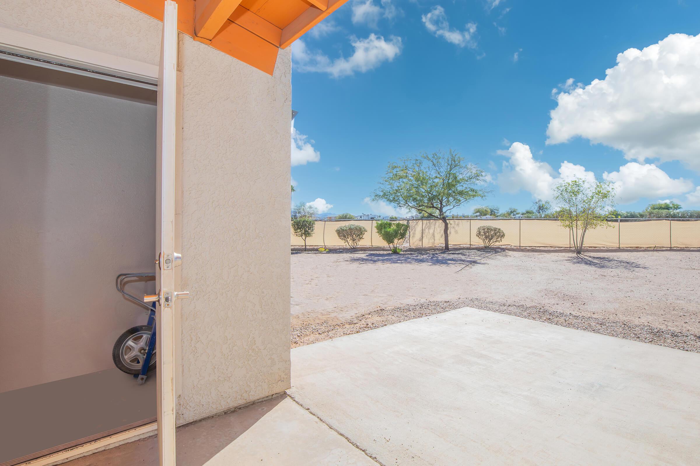 View from an open door leading outside to a sunny landscape. The scene shows a clear blue sky with fluffy white clouds, a flat, dry area with sparse vegetation, and a fence in the background. A partially visible cart or stroller is inside the doorframe.