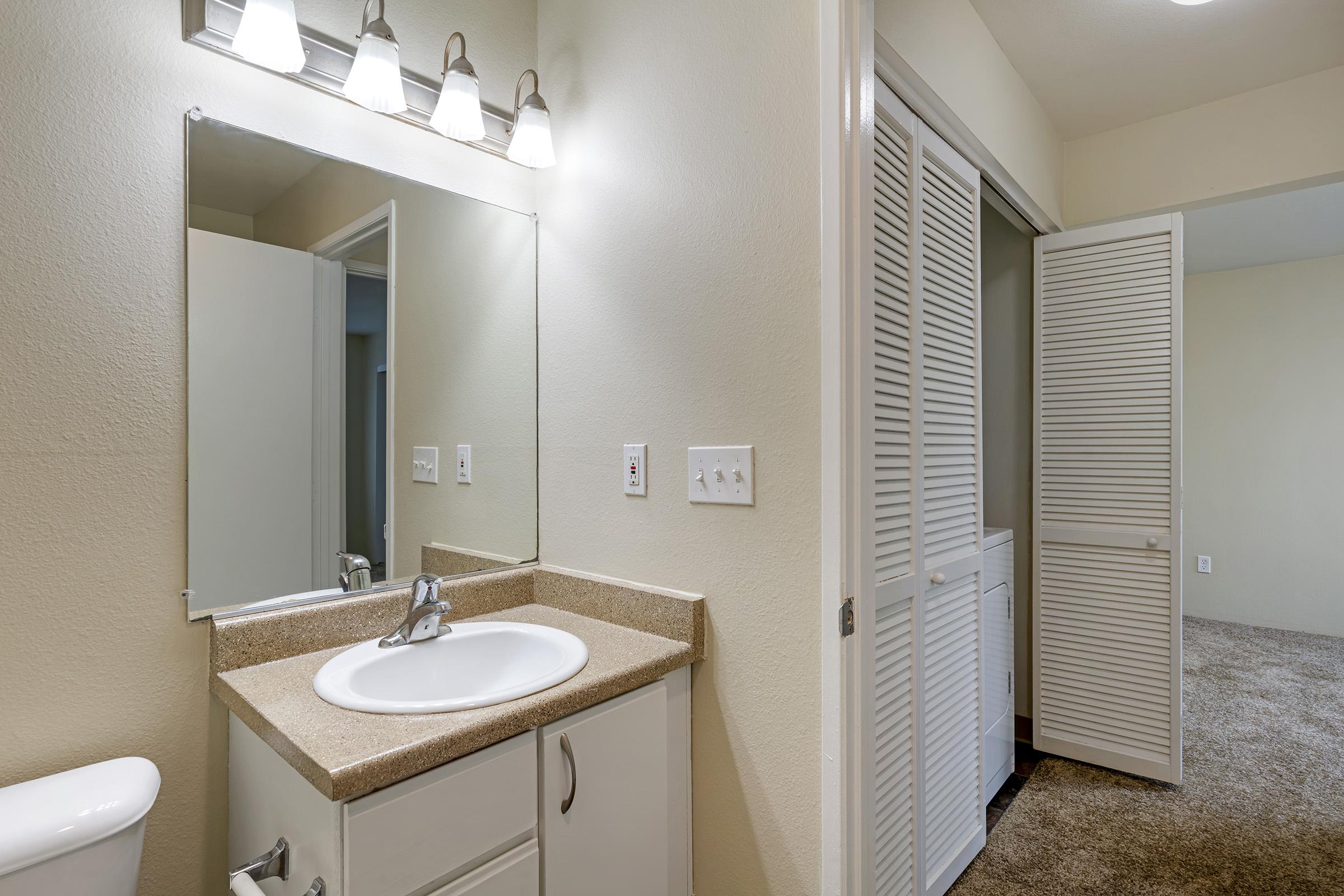 A well-lit bathroom featuring a countertop sink with a modern faucet, a large mirror above, and light fixtures. There are light beige walls and a door leading to an adjacent room with a carpeted floor and closed folding doors, indicating ample storage space.
