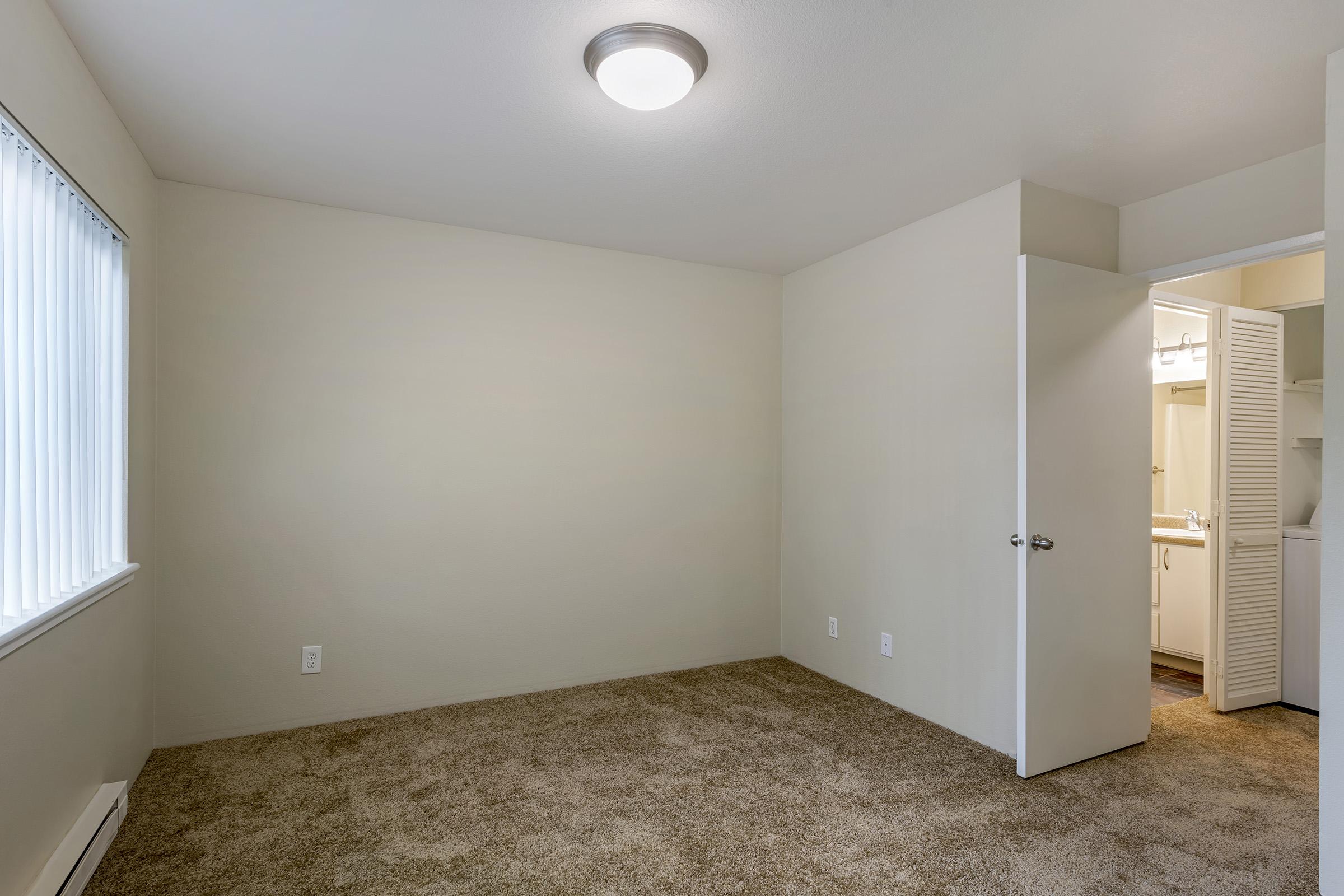 A bare room with light beige walls and carpet. A ceiling light is visible. There are vertical blinds on a window to the left. A partially open door on the right leads to a bathroom area, indicating a closet space. The overall aesthetic is simple and clean.