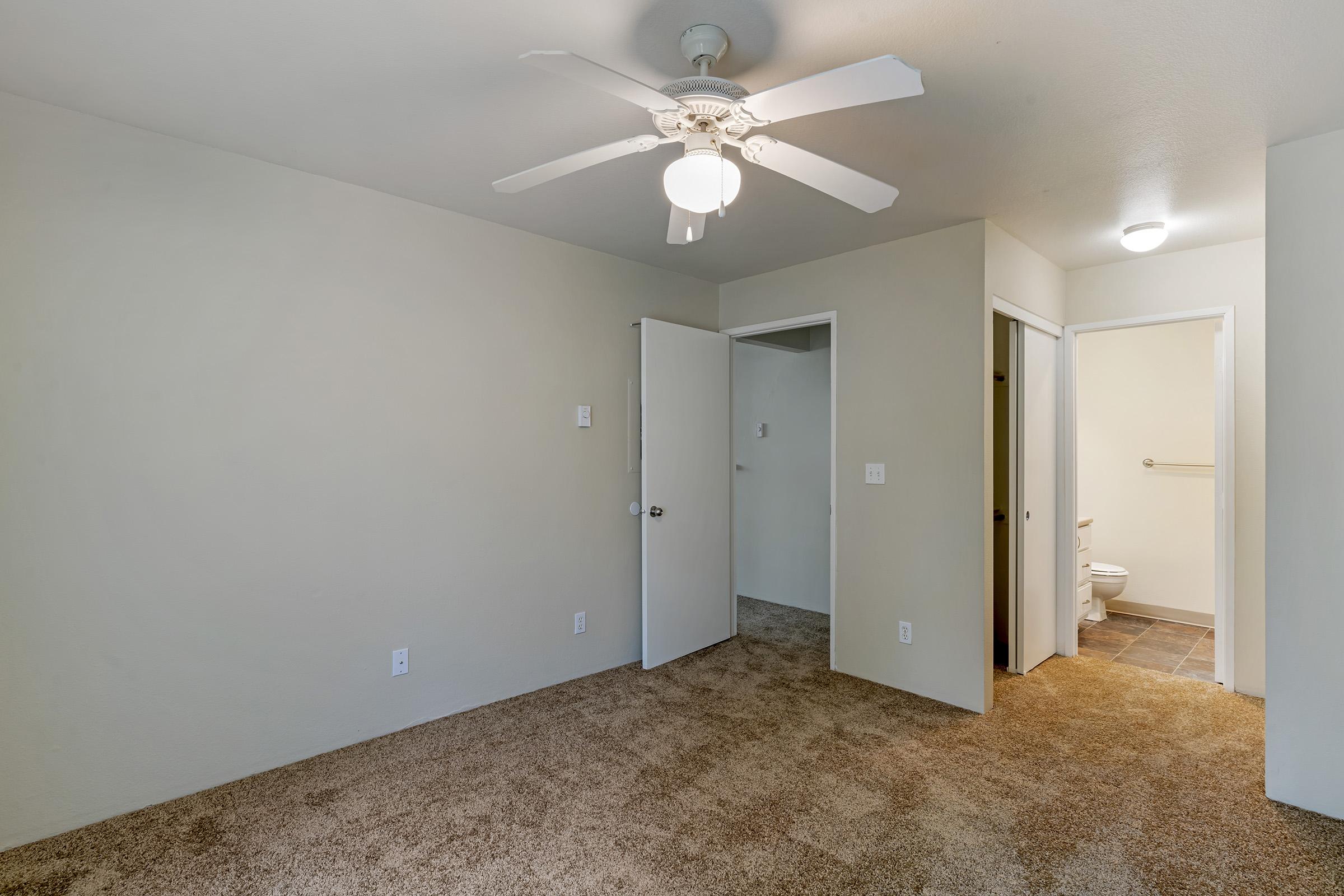 A view of a spacious, empty room featuring beige walls and a light brown carpet. A ceiling fan is installed, and there are two doors leading to other rooms or a bathroom. The room is well-lit, with a light fixture on the ceiling, and has minimal furnishings, showcasing its potential for customization.