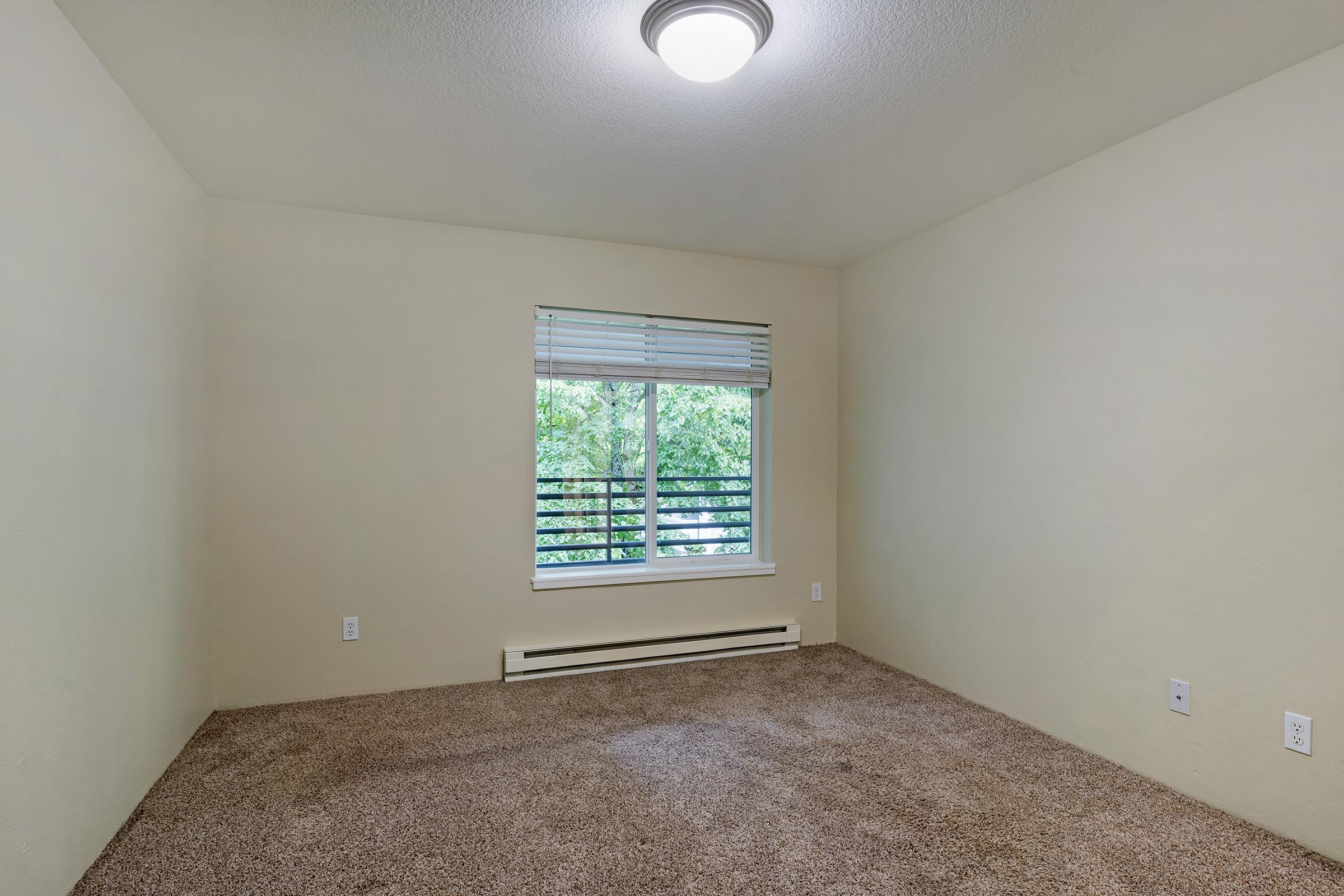 A vacant room with beige walls and a carpeted floor. There's a window with white blinds allowing natural light, and a ceiling light fixture. The room appears clean and spacious, with no furniture present.
