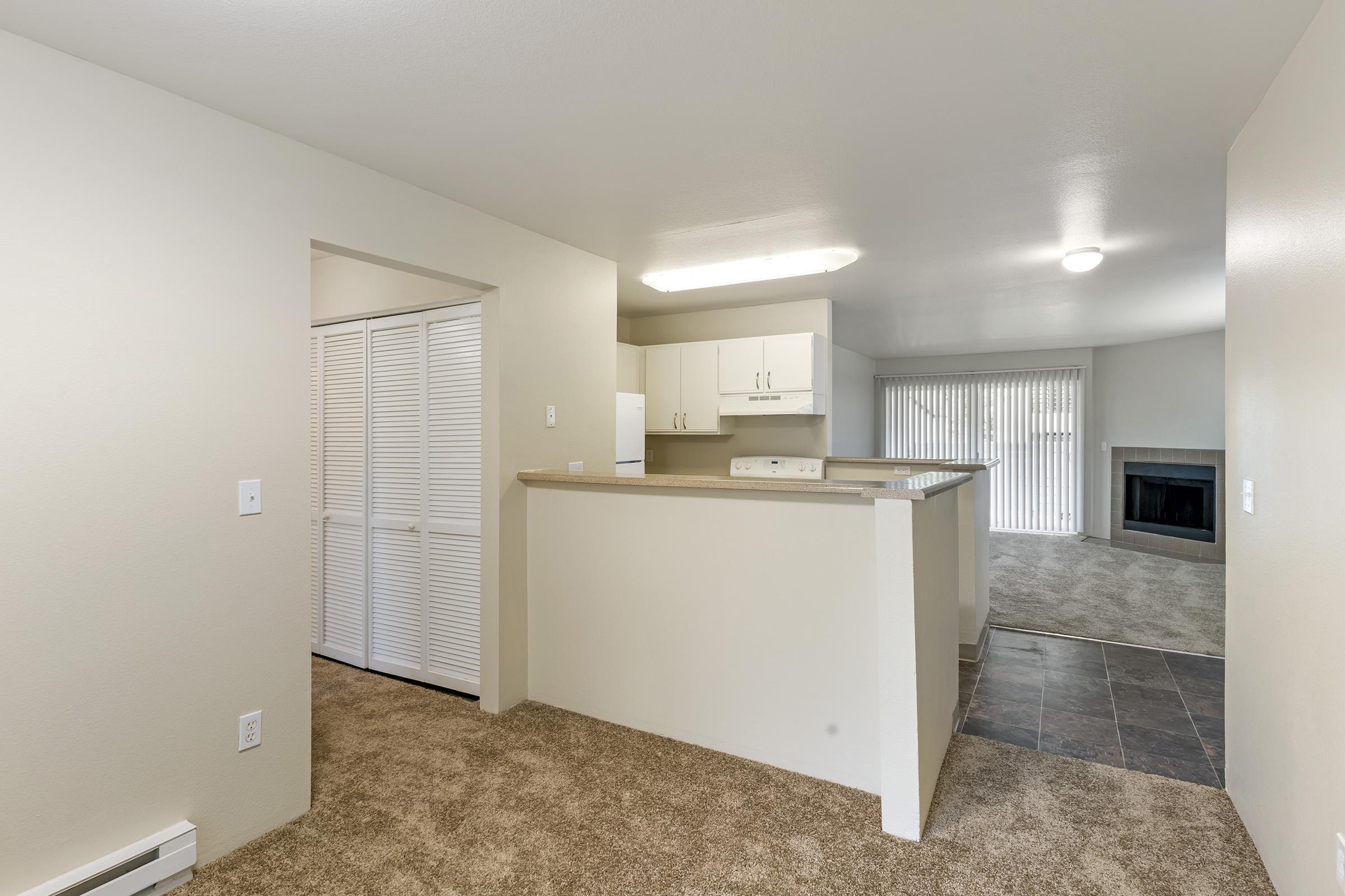 Interior view of an apartment featuring a kitchen with white cabinets and a breakfast bar. The living area is carpeted, leading to sliding glass doors with vertical blinds and a fireplace. A hallway is visible with closet doors. Neutral-colored walls and flooring create a spacious ambiance.