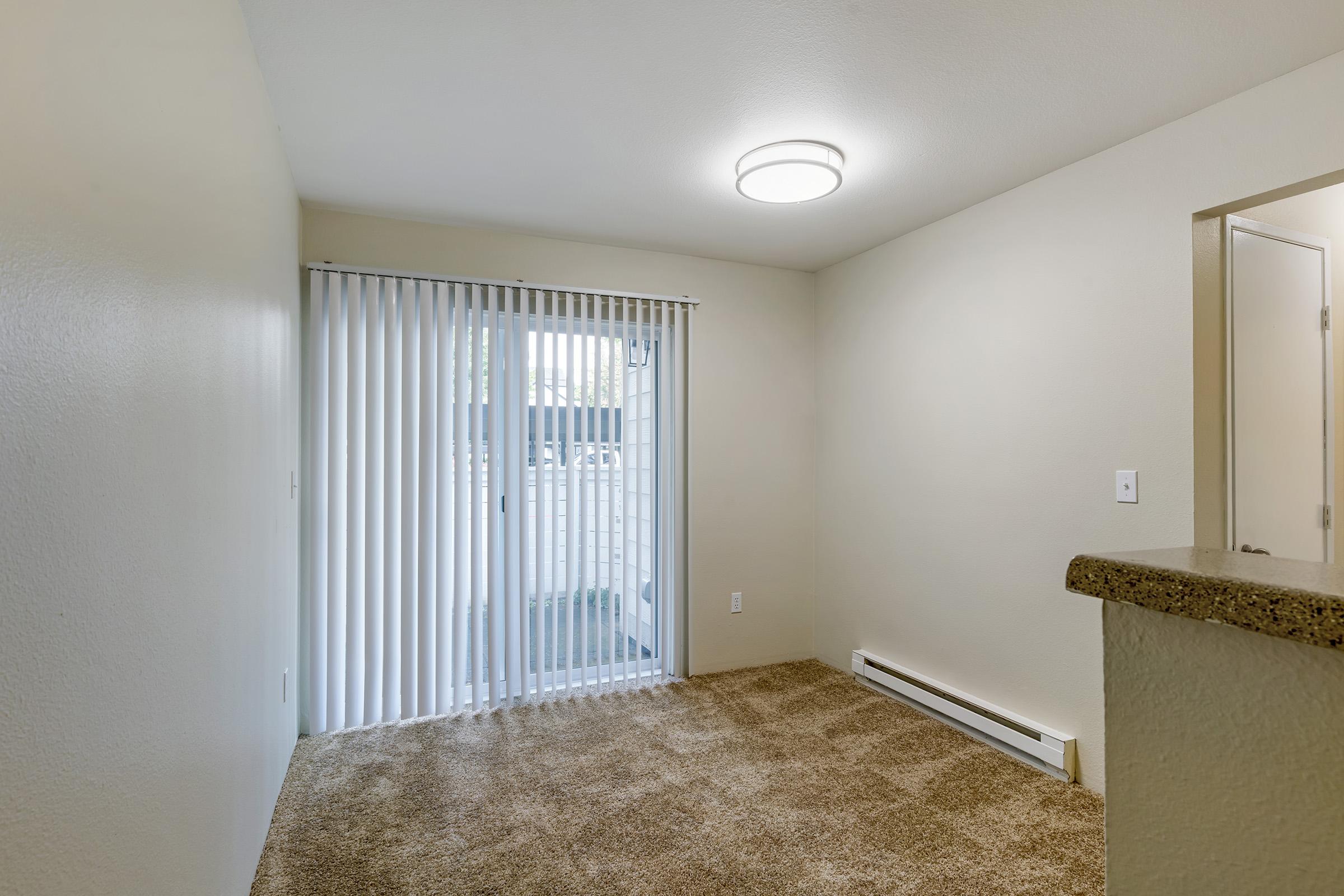 A well-lit, empty living space featuring beige walls and carpet. A sliding glass door with vertical blinds leads to a small outdoor area. There is a modern ceiling light fixture and a counter visible in the foreground, suggesting an open layout. The overall atmosphere is clean and inviting.