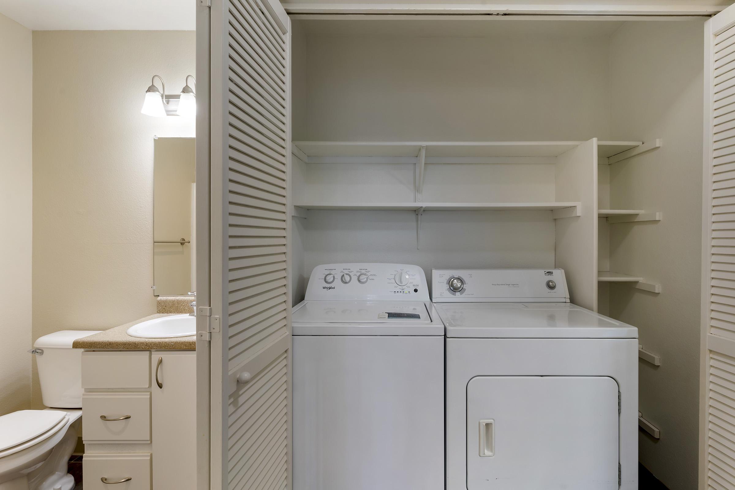 A laundry area featuring a washing machine, dryer, and storage shelves. The space is tidy with a light-colored wall and includes a bathroom vanity with a mirror and a toilet in the background. The shelves provide additional storage options in the room.