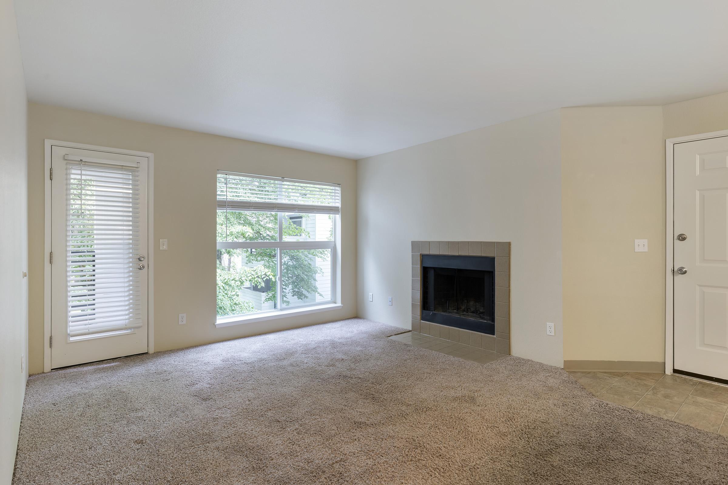 A spacious living room with light beige walls and carpeted flooring. There's a large window with blinds allowing natural light, and a fireplace with a simple stone surround. A door leads outside, and another door is visible, likely leading to another room or entrance.