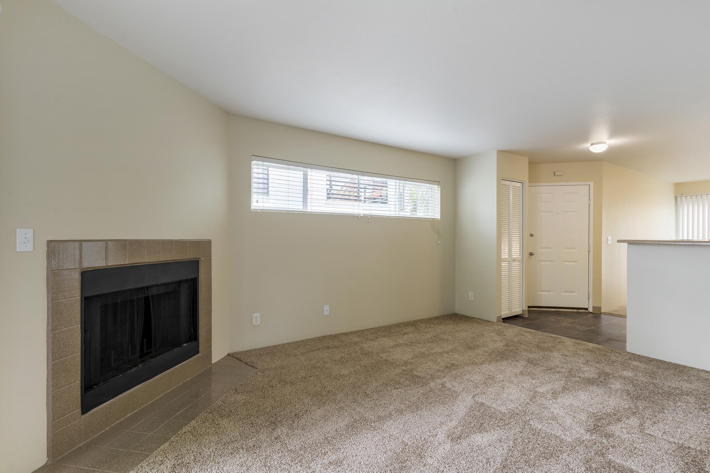 Interior shot of a living space featuring a beige carpet, a window with blinds allowing natural light, a fireplace with a tile surround, and a doorway leading to an entrance. The walls are painted a light color, creating a spacious and inviting atmosphere.