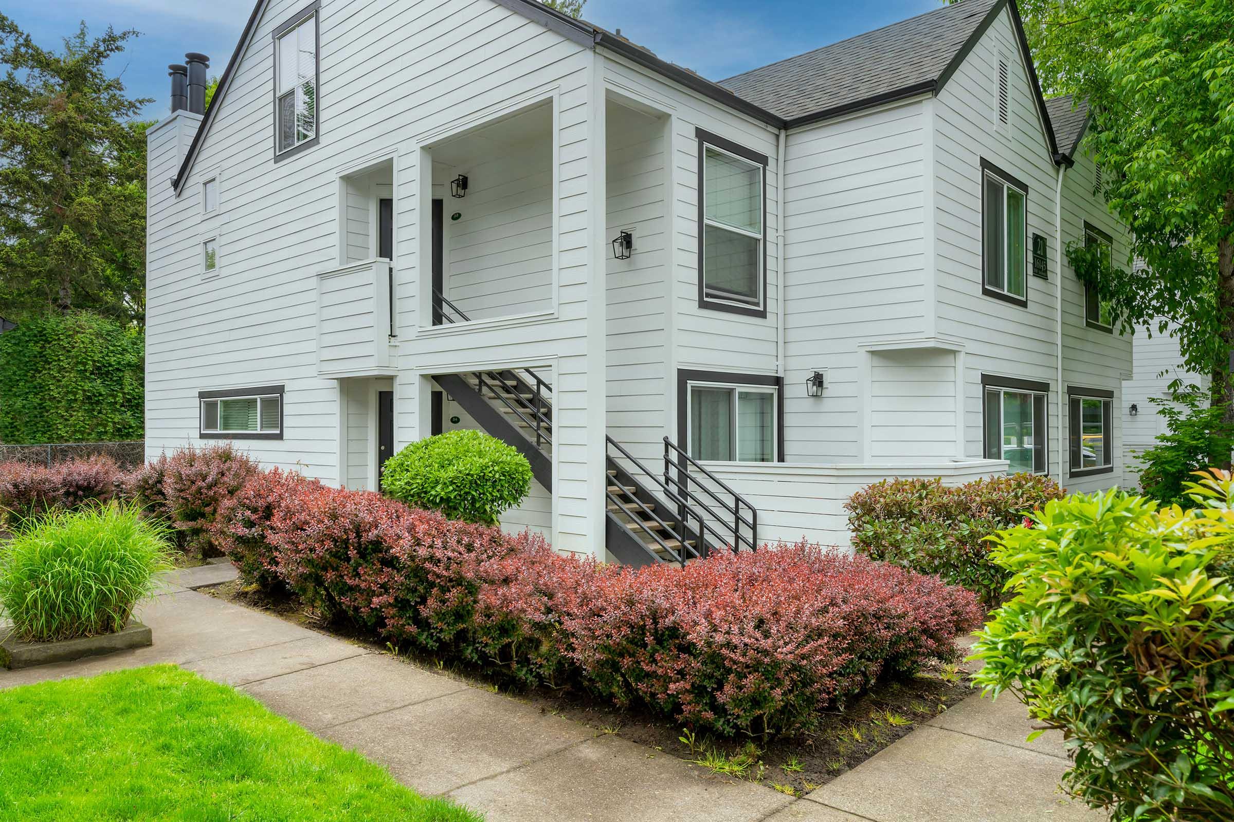A modern white two-story apartment building surrounded by greenery. The building features large windows, a staircase leading to the second floor, and neatly trimmed bushes in vibrant colors. The pathway is paved with concrete, and the lawn is lush and well-maintained.