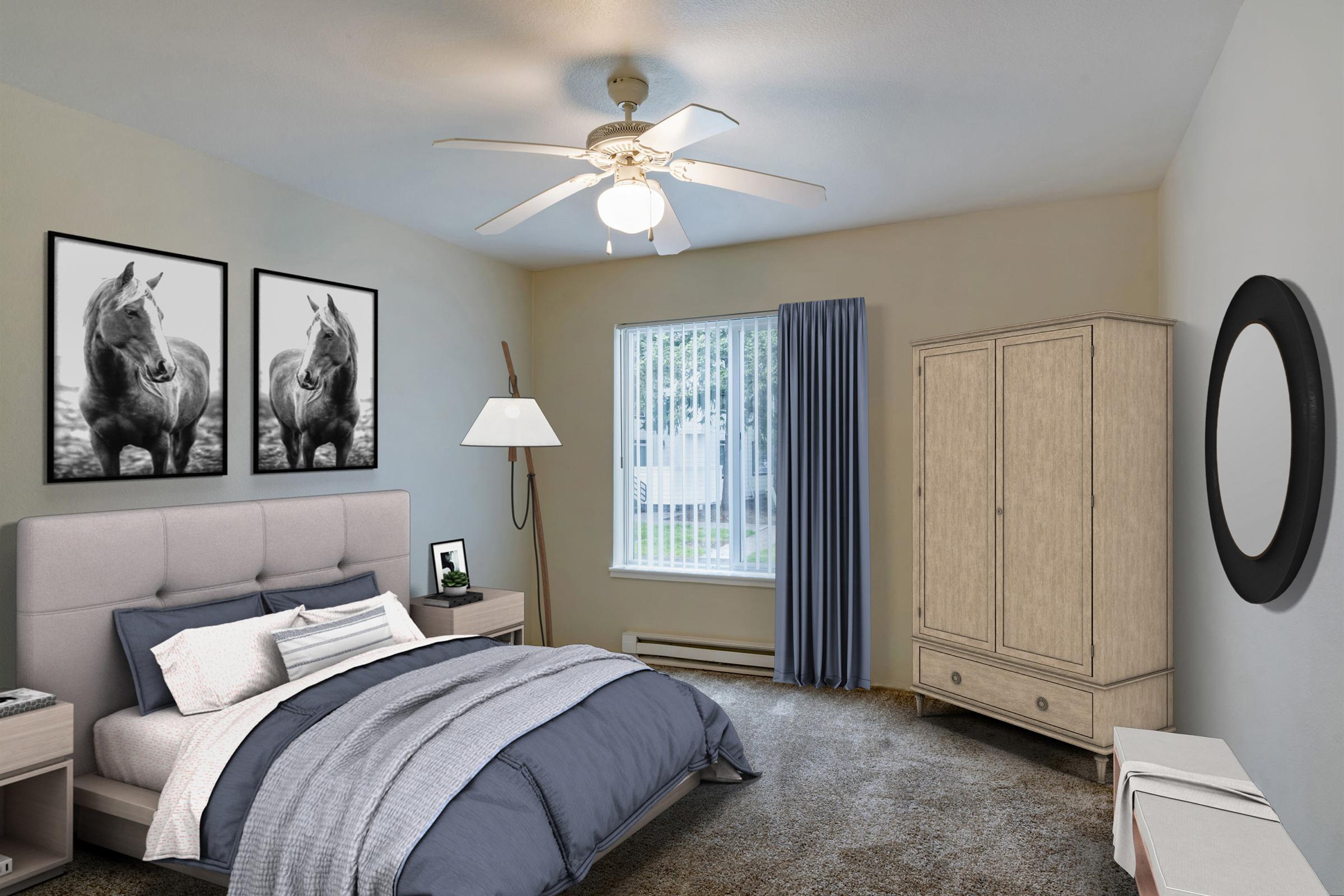 A modern bedroom featuring a gray upholstered bed with layered bedding, two horse-themed black and white framed pictures on the wall, a ceiling fan, a nightstand with a lamp, a curtain-covered window, a tall wardrobe, and a round mirror, all set against a neutral-colored wall.
