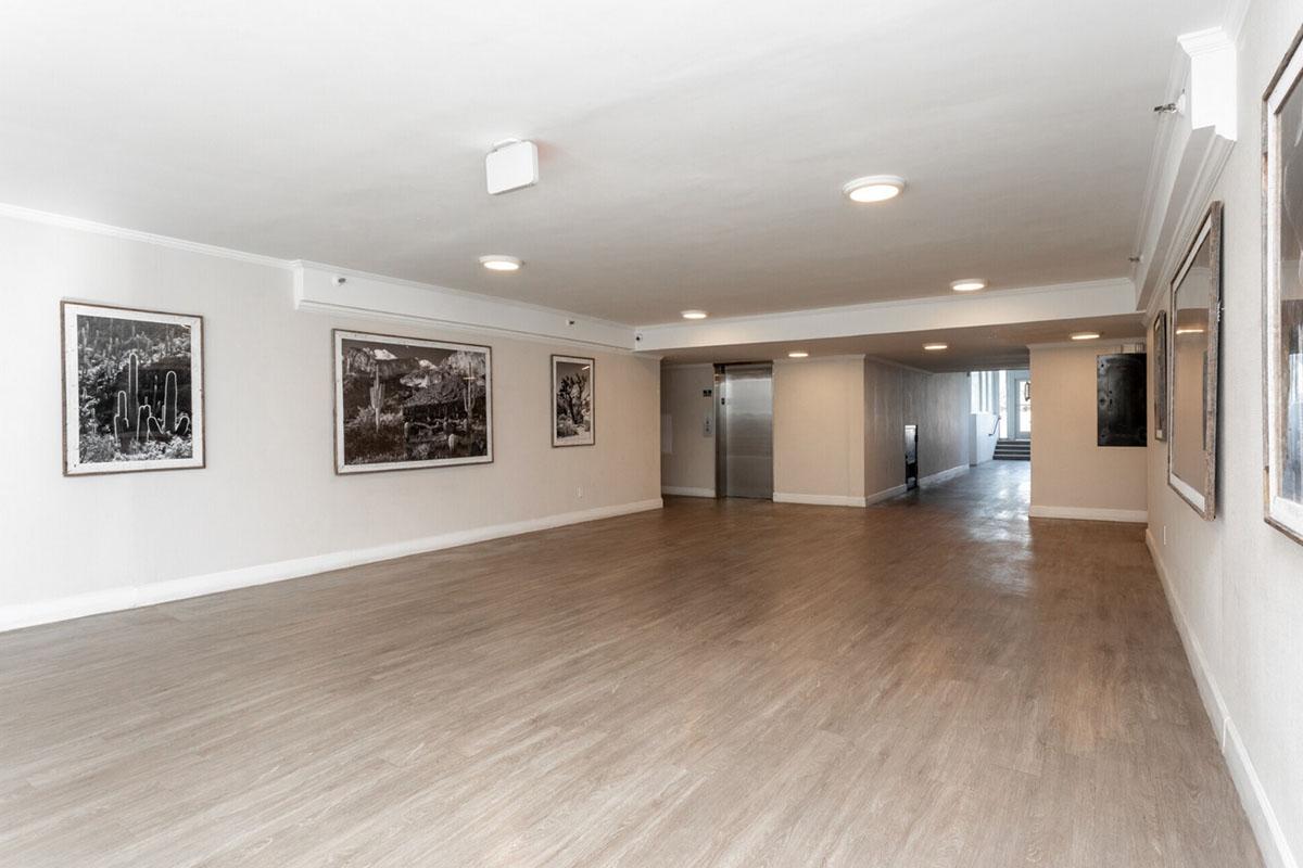 A spacious hallway with smooth light-colored flooring and white walls. Framed black-and-white photographs are hung on the walls. At the far end, there is an elevator and additional room access. The area is well-lit with overhead lights. The ambiance is modern and minimalistic.