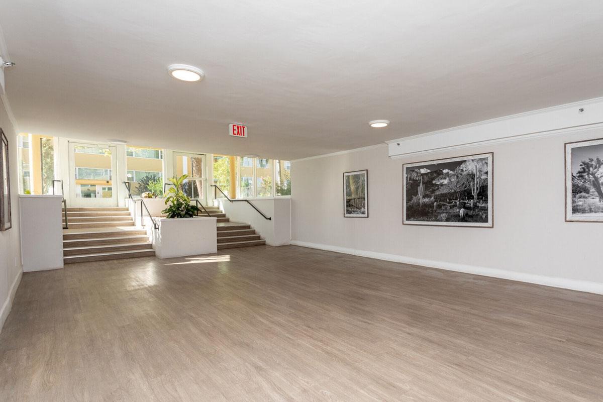A spacious, well-lit lobby area featuring light-colored walls and wooden flooring. There are large windows allowing natural light, and a staircase leading to an upper level. Framed black and white photographs adorn the walls, and a few green plants add a touch of nature to the space.