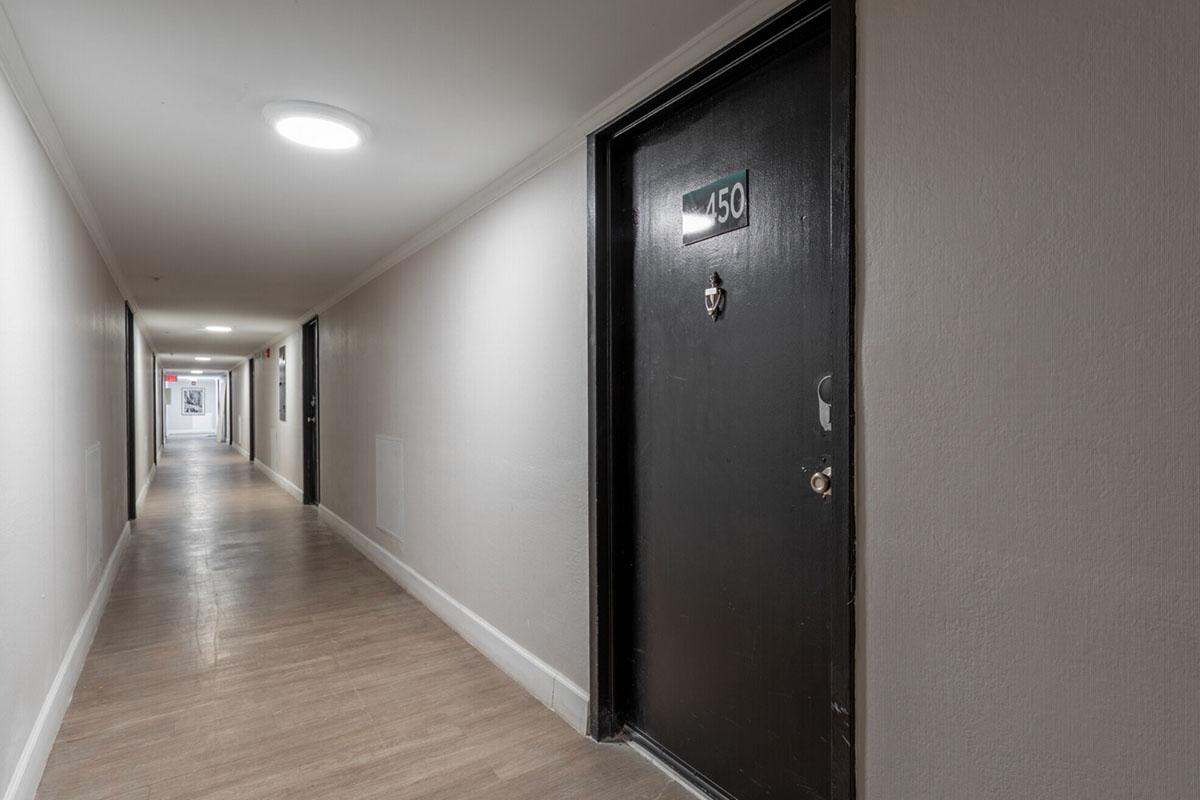 A long, well-lit hallway with light gray walls and wooden flooring. Doors line both sides, with the focus on a black door labeled "450." Soft lighting from overhead fixtures creates a welcoming atmosphere, leading to a distant view of the end of the corridor.