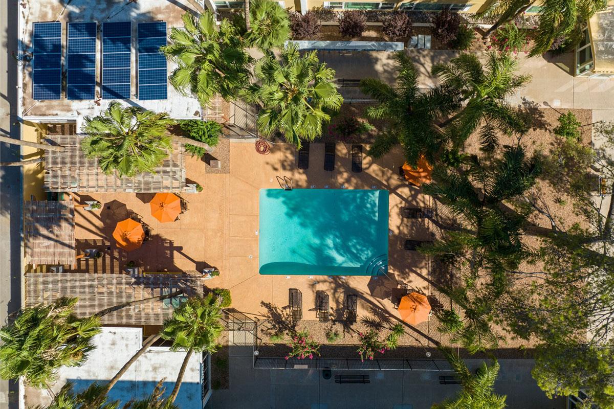 Aerial view of a resort-style area featuring a turquoise swimming pool surrounded by lounge chairs and orange umbrellas. Lush palm trees line the space, with solar panels visible on one rooftop. The scene is set in a sunny, landscaped environment, highlighting a relaxing outdoor atmosphere.