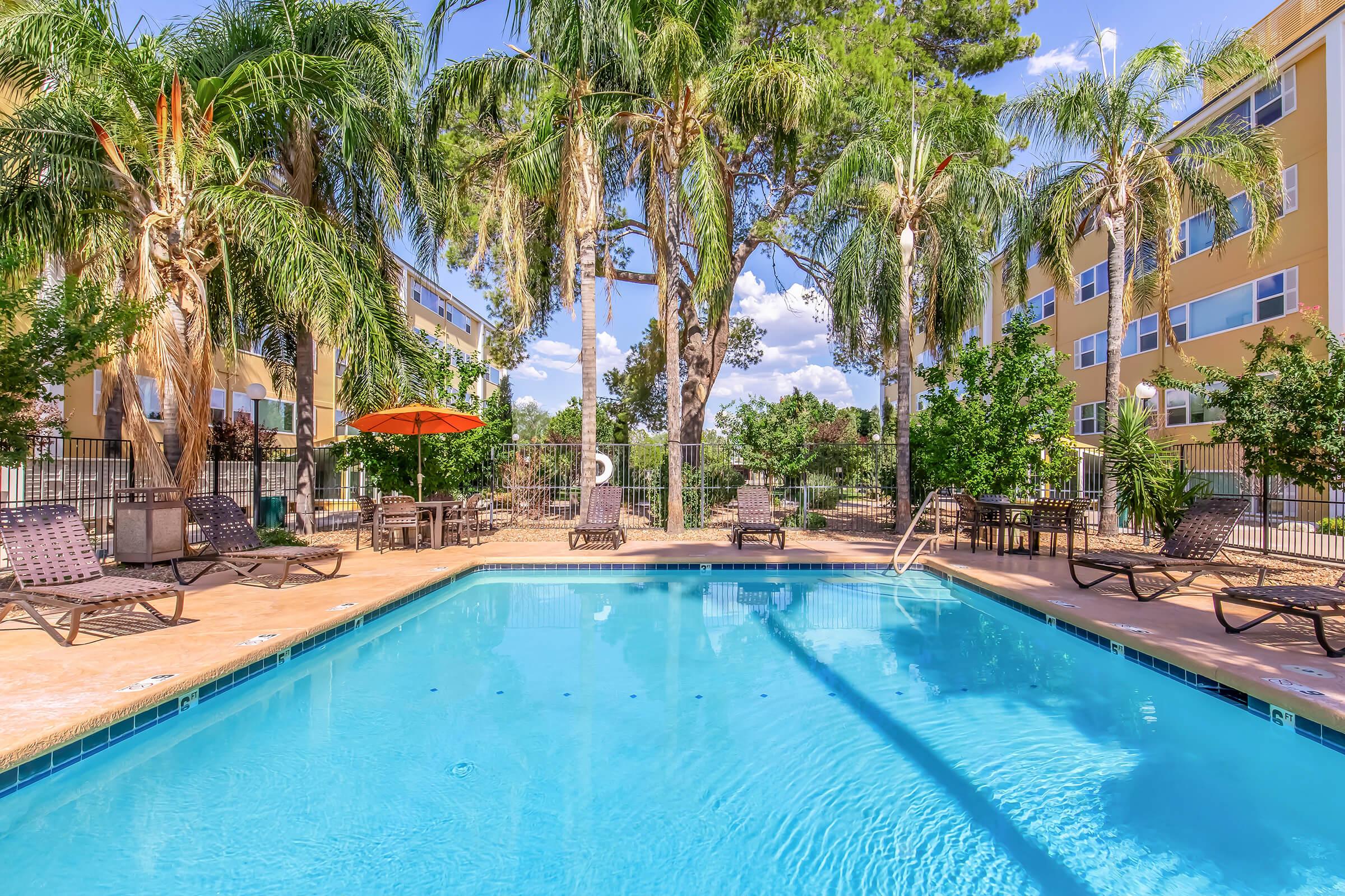 A bright outdoor swimming pool surrounded by palm trees and lounge chairs. An orange umbrella provides shade near the poolside. In the background, there are apartment buildings and a clear blue sky with a few clouds. The setting is inviting and perfect for relaxation.