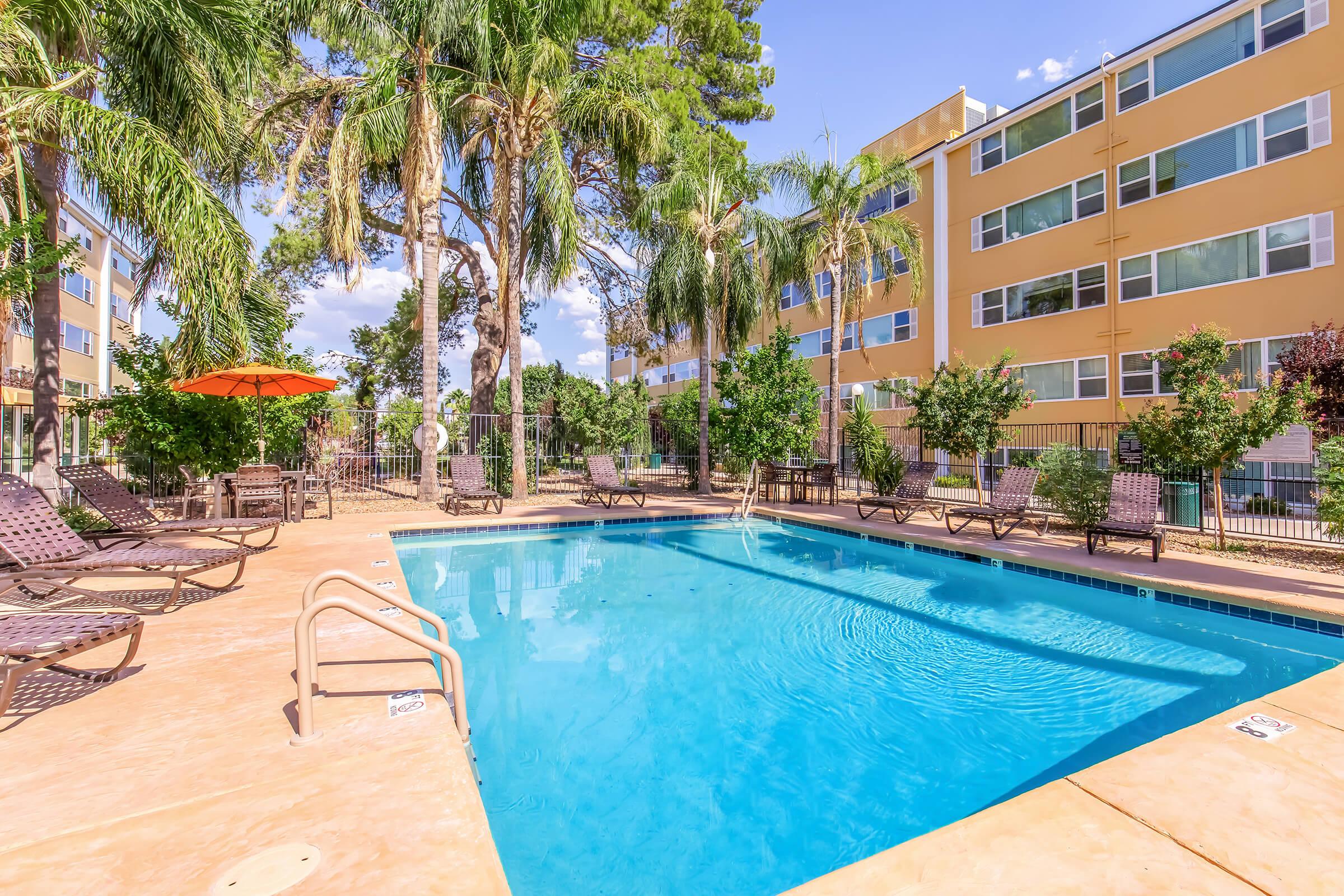 A sunny outdoor pool area featuring a clear blue swimming pool surrounded by lounge chairs and palm trees. An orange umbrella provides shade, with a backdrop of a multi-story yellow building and a clear blue sky. The serene setting invites relaxation and enjoyment.