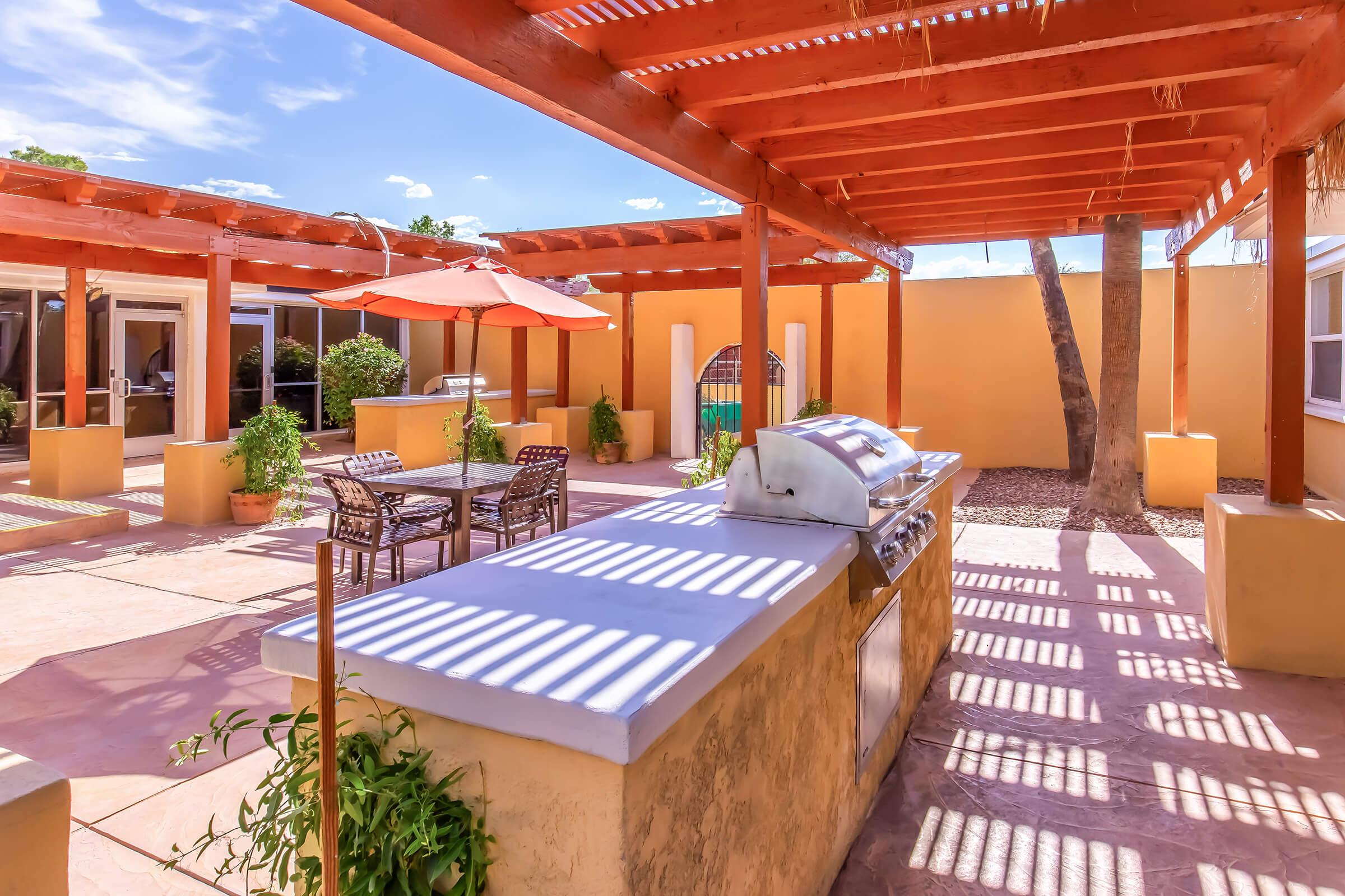 A spacious outdoor patio featuring a built-in BBQ grill, surrounded by shaded pergolas. There are tables and chairs for seating, along with potted plants. The warm-colored walls and sunlight create a welcoming atmosphere, perfect for gatherings or relaxation.