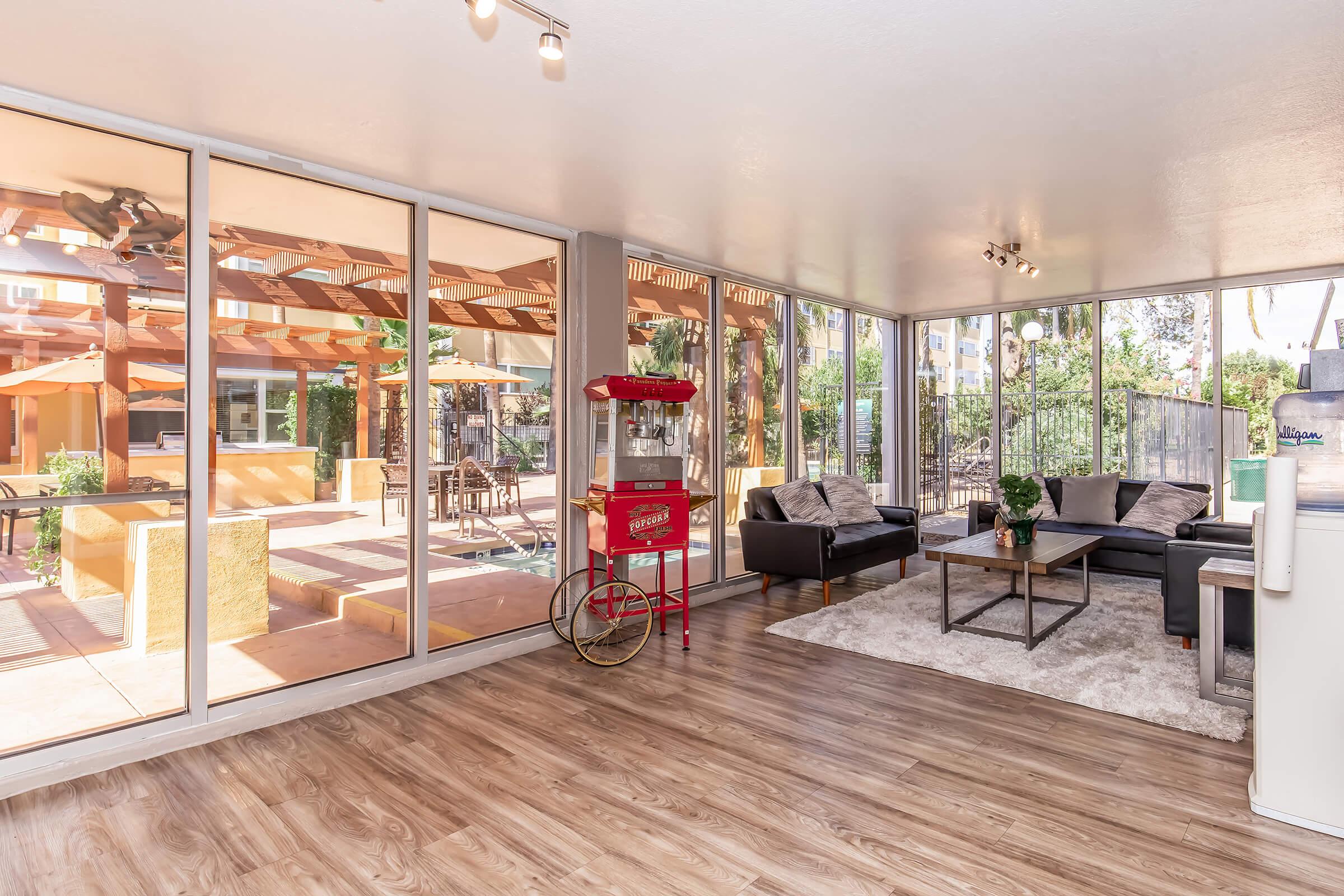 Bright and inviting sunroom featuring large glass windows that showcase a patio area. Inside, there are modern couches, a coffee table, and a vintage popcorn machine, adding a playful touch. The warm wooden floor complements the overall cozy ambiance, creating a perfect space for relaxation and entertainment.
