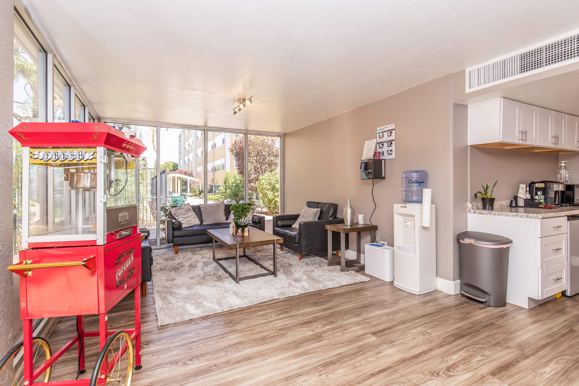 A modern living area featuring a comfortable black sofa set, a wooden coffee table, and a stylish rug. Nearby, a red popcorn machine adds a playful touch. A water cooler and a trash can are positioned against the wall, with large windows showcasing greenery outside, creating a bright and inviting atmosphere.
