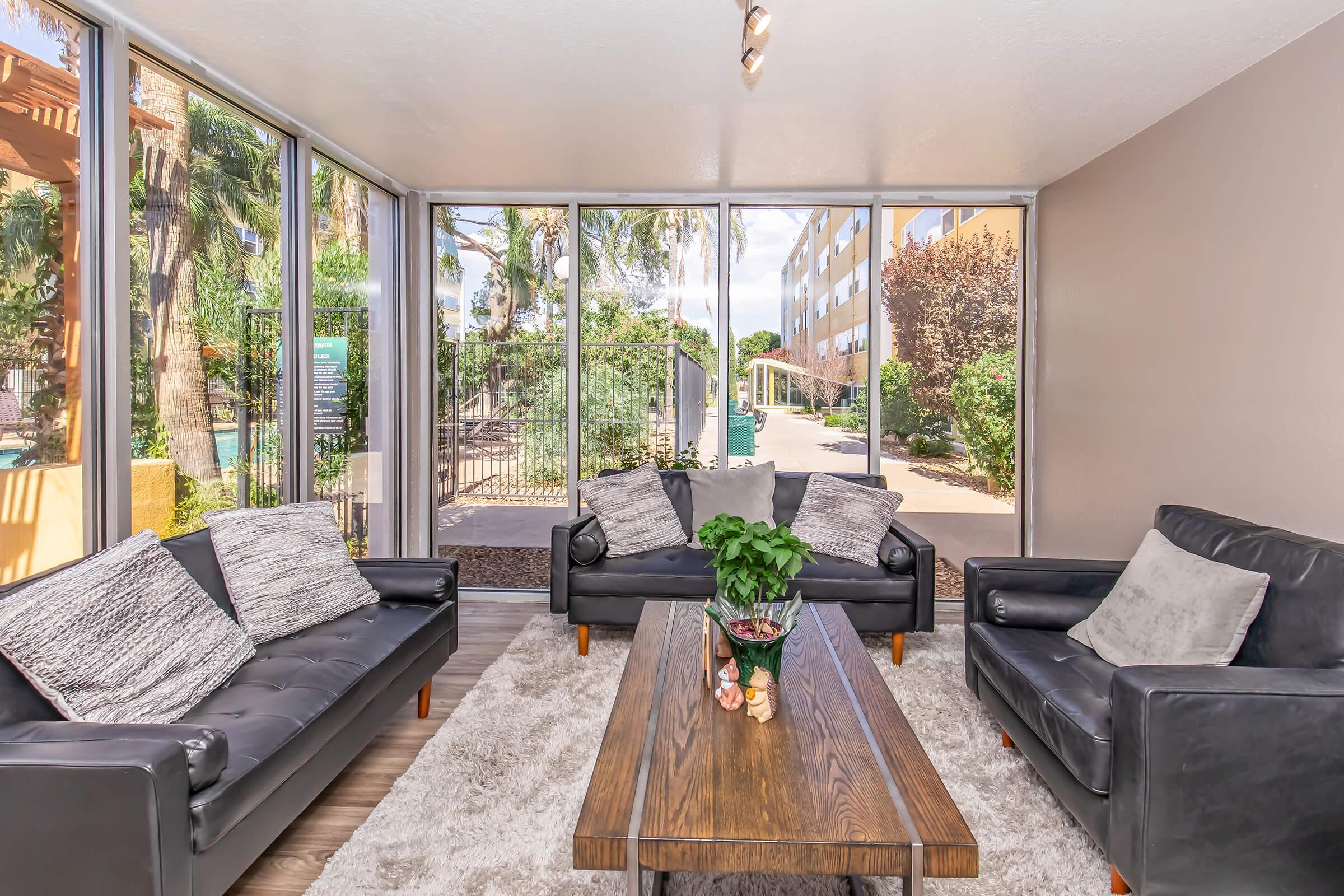 A modern living room featuring two black leather sofas with decorative pillows, a wooden coffee table with a plant centerpiece, and a plush area rug. Large windows let in natural light, offering a view of outdoor greenery and a pool area. The walls are light-colored, creating a bright and inviting atmosphere.