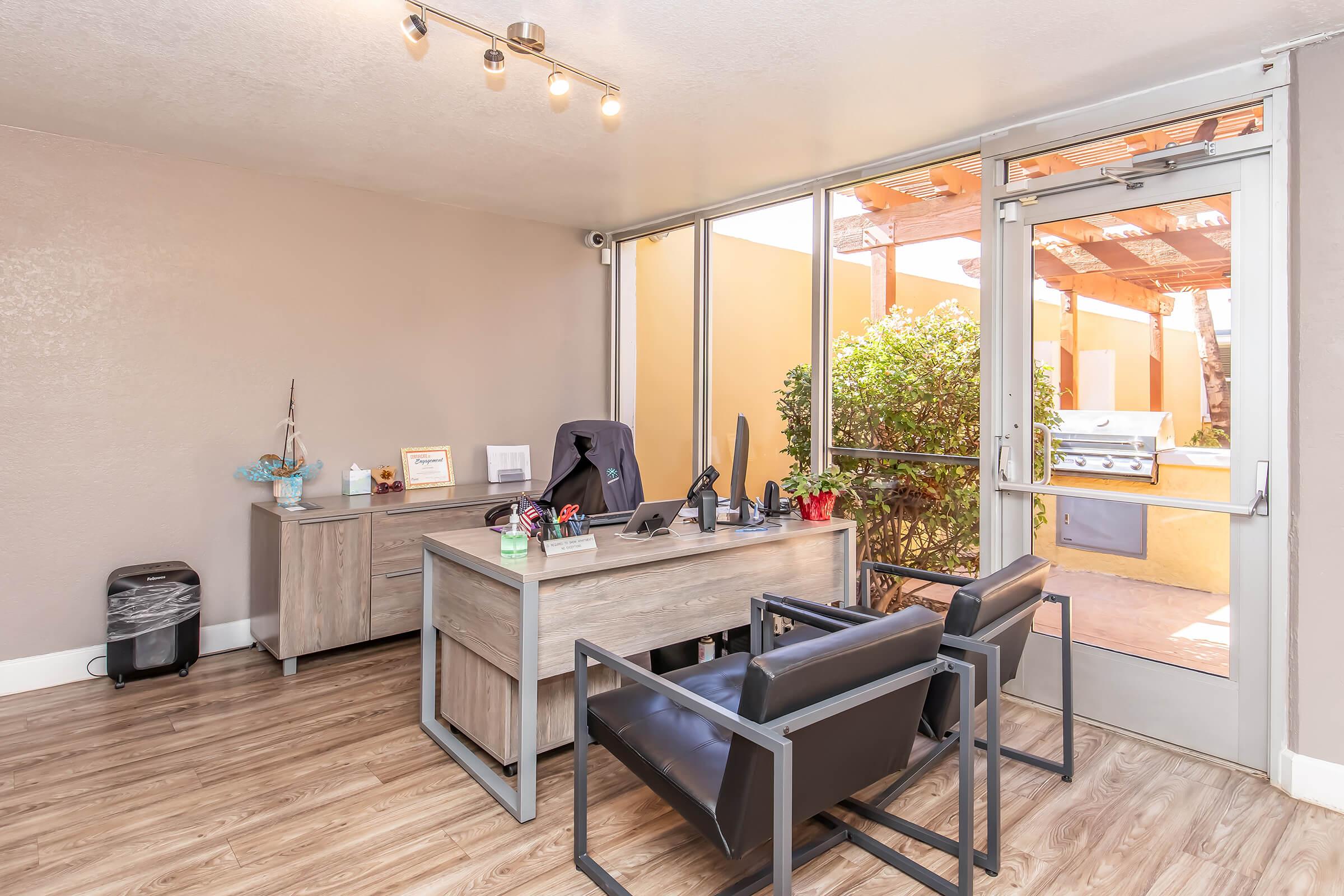 Modern office space featuring a wooden desk with office supplies, a computer, and a chair. Two black chairs are positioned in front. Large windows allow natural light to illuminate the room, revealing a view of greenery outside. The decor is minimalistic and professional, with warm wood flooring.