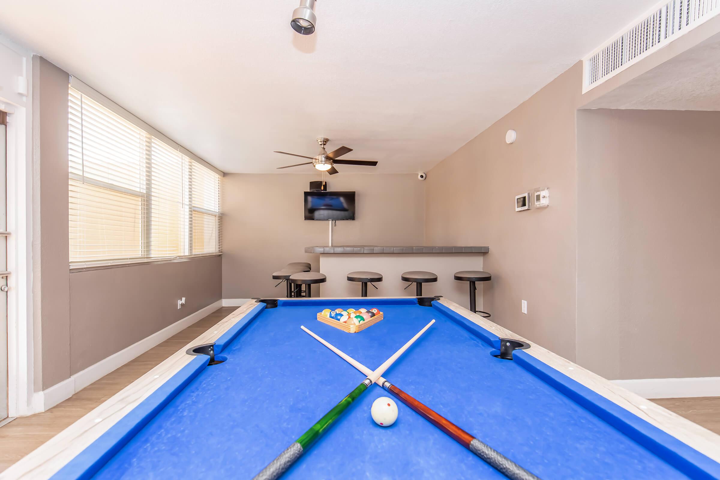A blue billiards table with cue sticks crossed over it, set up for a game. In the background, there are bar stools and a flat-screen TV mounted on the wall. Large windows allow natural light to fill the room, creating a warm and inviting atmosphere.