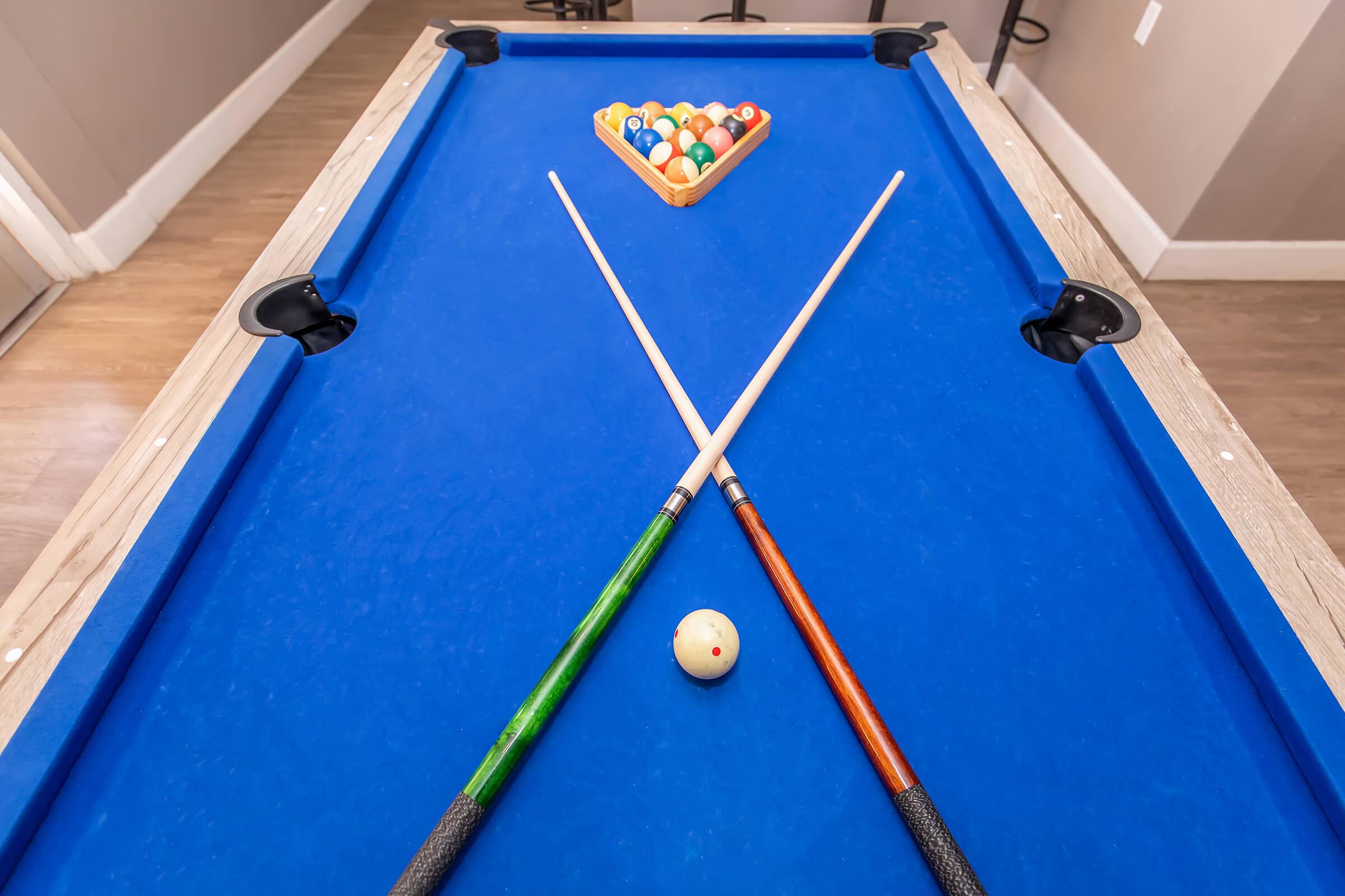 A blue billiard table with two cue sticks crossed over a white billiard ball. In the background, a triangle rack holds colorful billiard balls, indicating a game in progress or about to start. The setting is indoors, with neutral-colored walls and flooring.